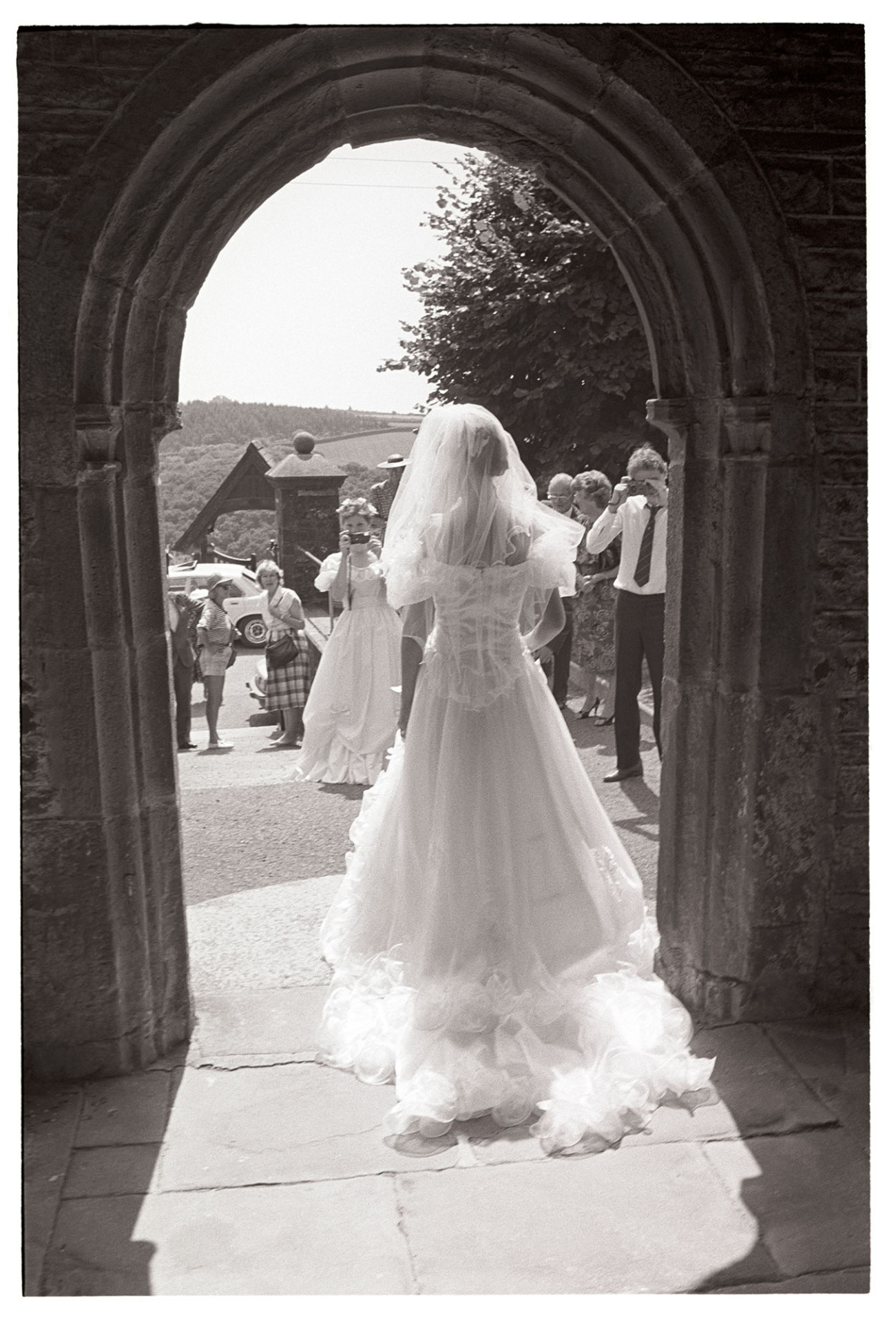 Wedding, bride at church door after wedding. 
[A bride posing at the door of Chulmleigh Church while guests take photographs from outside. The image is taken from inside Chulmleigh Church.]
