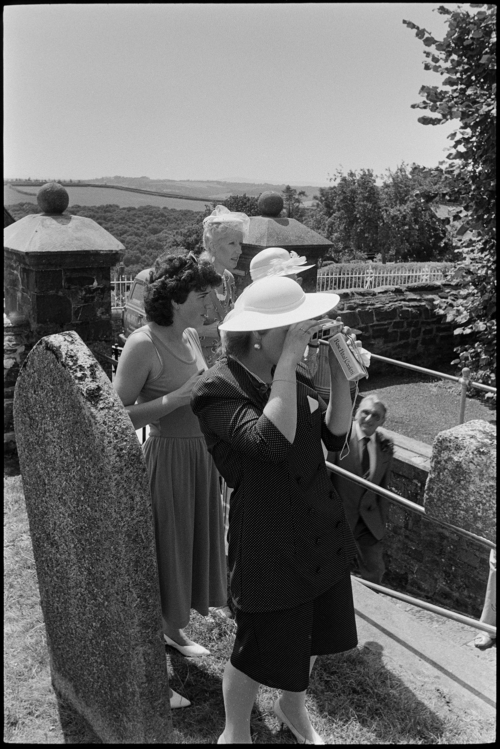 Wedding spectators, bride, groom, bridesmaids being photographed Photographer changing film.
[A group of four women standing in the churchyard of the Church of St Mary Magdalene, Chulmleigh taking photographs at a wedding. The women are wearing hats.]