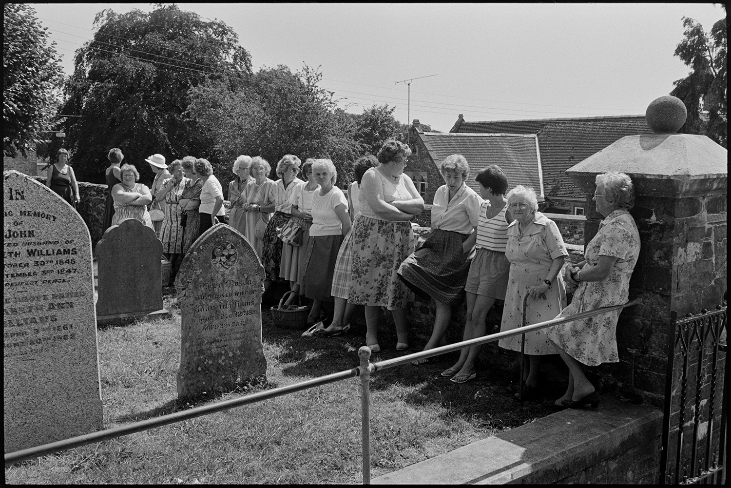 Wedding spectators, bride, groom, bridesmaids being photographed Photographer changing film.
[A line of women leaning against the wall of the churchyard of the Church of St Mary Magdalene, Chulmleigh waiting to see a wedding. Chulmleigh School can be seen in the background.]