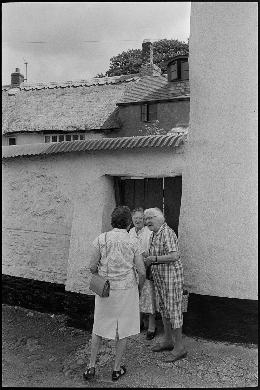 Fair, auction of sheep, farmers and crowds of spectators.
[Three women talking in a street in Chulmleigh on the day of Chulmleigh Fair. A thatched building can be seen in the background.]