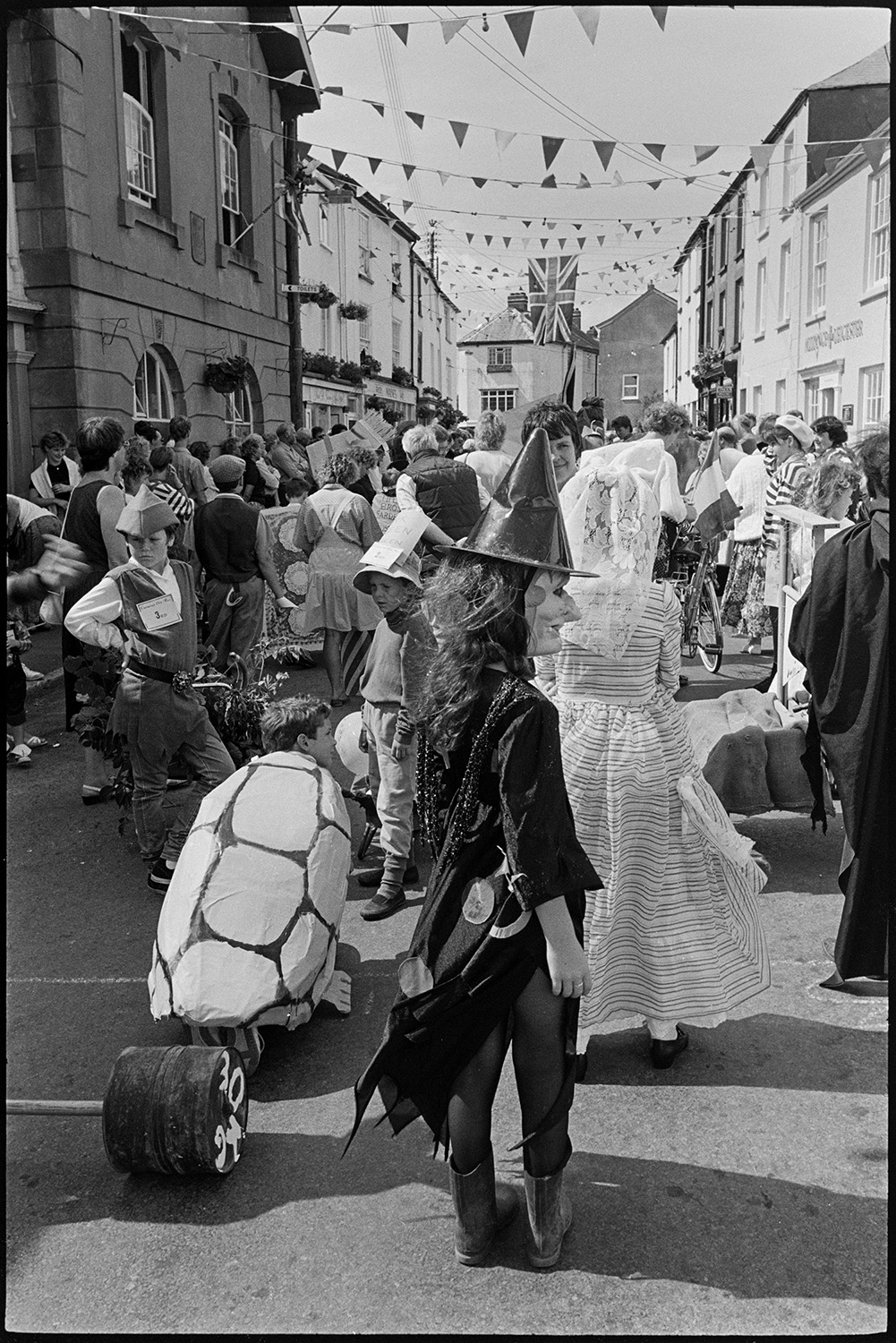 Fair, display of flowers and produce, fancy dress parade in street. 
[People in fancy dress gathered in a street decorated with bunting at Chulmleigh Fair. A person in the foreground is dressed as a witch and a child nearby is in a homemade cannon.]