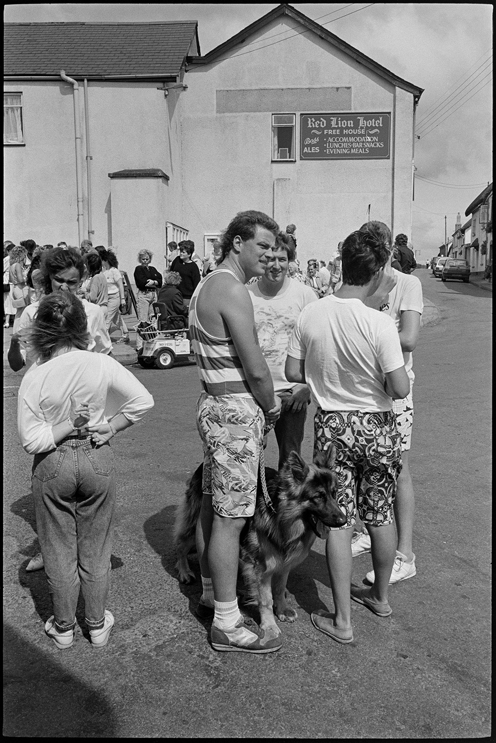 Fair, display of flowers and produce, fancy dress parade in street. 
[Men and women gathered in the street outside the Red Lion Hotel at Chulmleigh Fair. A man is holding the lead of a German Shepherd dog in the foreground. Some of the young men are wearing patterned shorts.]