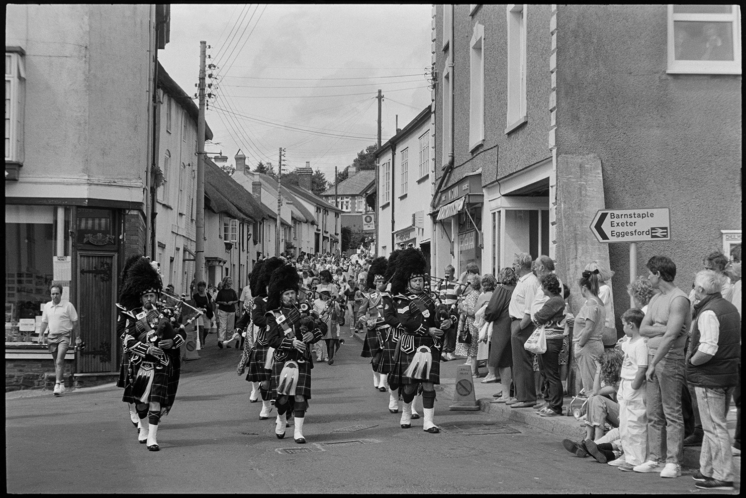 Fair, display of flowers and produce, fancy dress parade in street. 
[A crowd of people lining a street in Chulmleigh and watching  band of Scottish Pipers parading along the street at Chulmleigh Fair.]