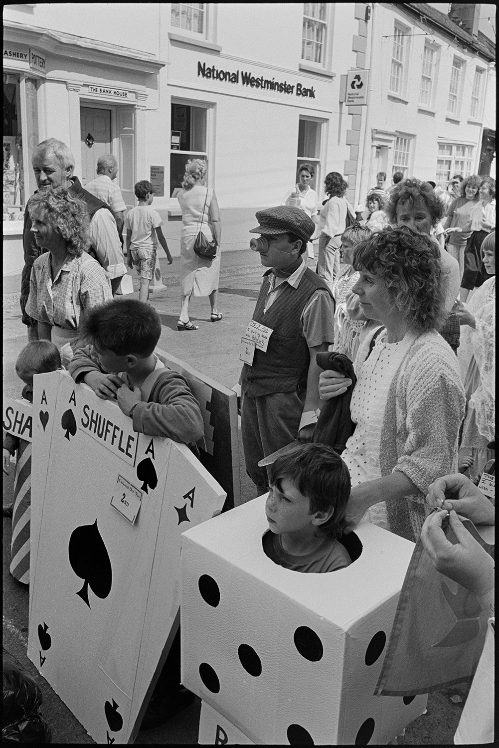 Fair, display of flowers and produce, fancy dress parade in street. 
[Children taking part in a fancy dress parade outside the National Westminster Bank at Chulmleigh Fair. They are dressed as a pack of cards and a die. Men and women are with them.]