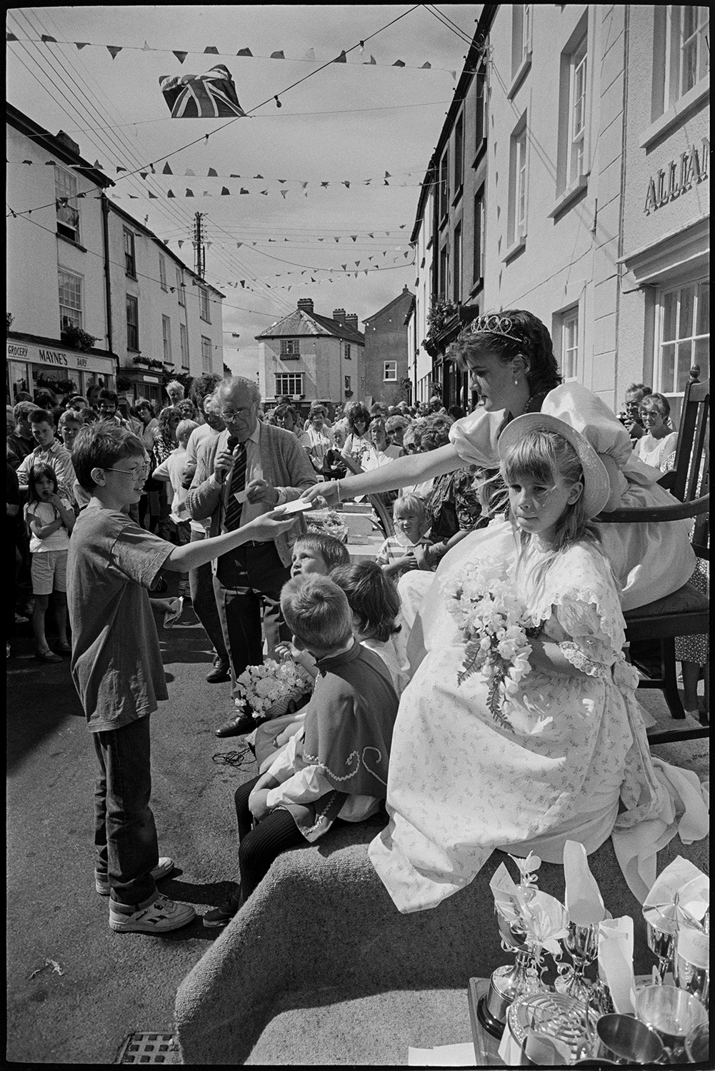 Fair, display of flowers and produce, fancy dress parade in street. 
[The Chulmleigh Fair Queen handing out  a prize to a boy in a street decorated with bunting in Chulmleigh. The Fair Queen and her attendants are sat on a podium in the street and a table of silver cups are visible in the foreground. A man is making announcements with a microphone and a crows is watching in the background.]