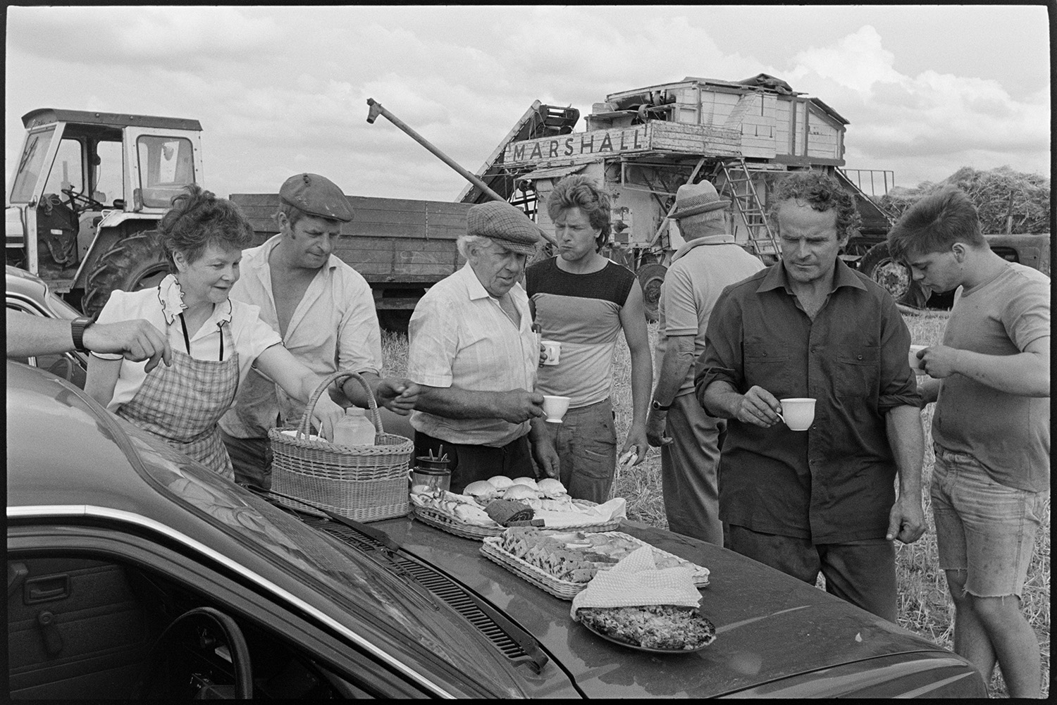 Reedcombers having tea break and working reed comber. 
[Members of the Down family and other men having a tea break with sandwiches and cake, from Reed combing in a field at Spittle Farm, Chulmleigh. Mrs Down is handing out items from a basket. A reed comber and tractor and trailer can be seen in the background.]