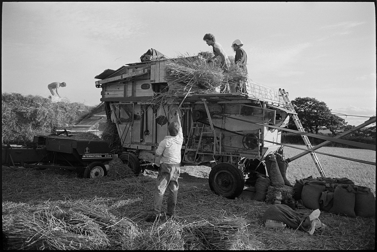 Reedcombers having tea break and working reed comber. 
[Men, including some from the Down family reedcoming in a field at Spittle Farm, Chulmleigh. A man in the foreground is lifting bundles of reed up to two men on the reed comber. One of the men is wearing a mask. Sacks of grain can be seen by the comber and in the background a man is stood on a trailer loaded with bundle of reed which have been through the reed comber.]