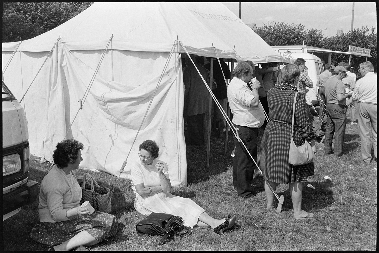 View over pens at sheep fair, farmers checking sheep, clerks at sale office in shed. 
[Men and women standing and sitting with drinks outside the refreshment tent at South Molton Sheep Fair.]