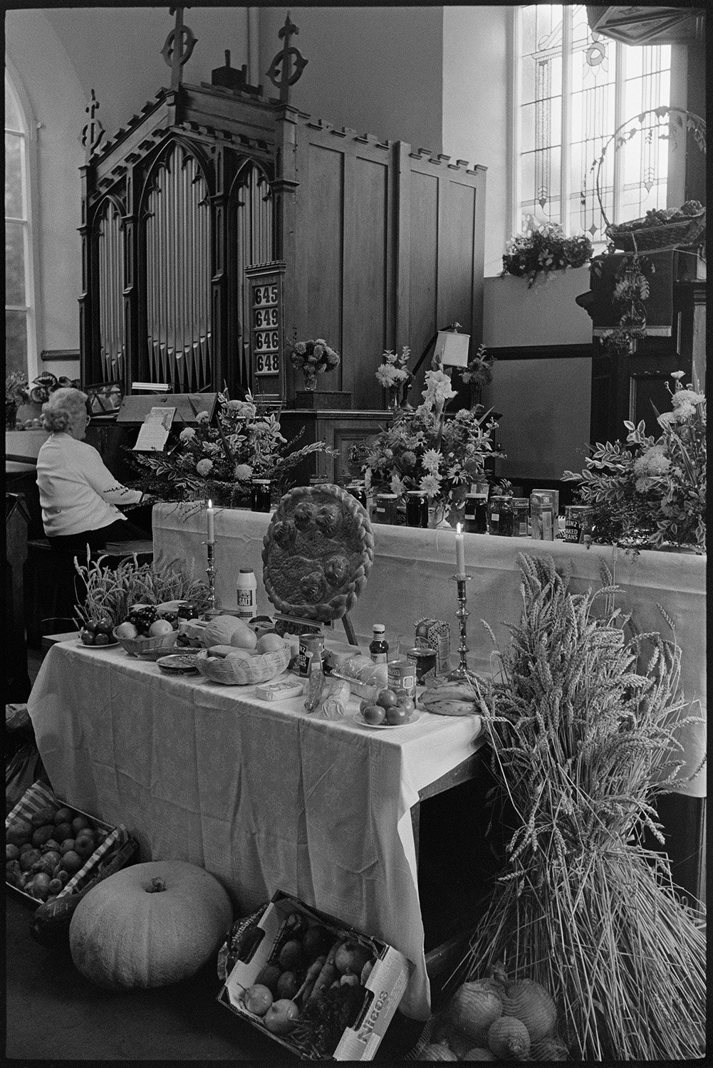 Harvest Festival display in Chapel, bread, produce and fruit. 
[A harvest festival display at the altar in Chulmleigh Congregational Chapel including sheaves of corn, swedes, jars of jam, tinned food, bananas and flower arrangements. A woman can be seen playing the organ in the background.]