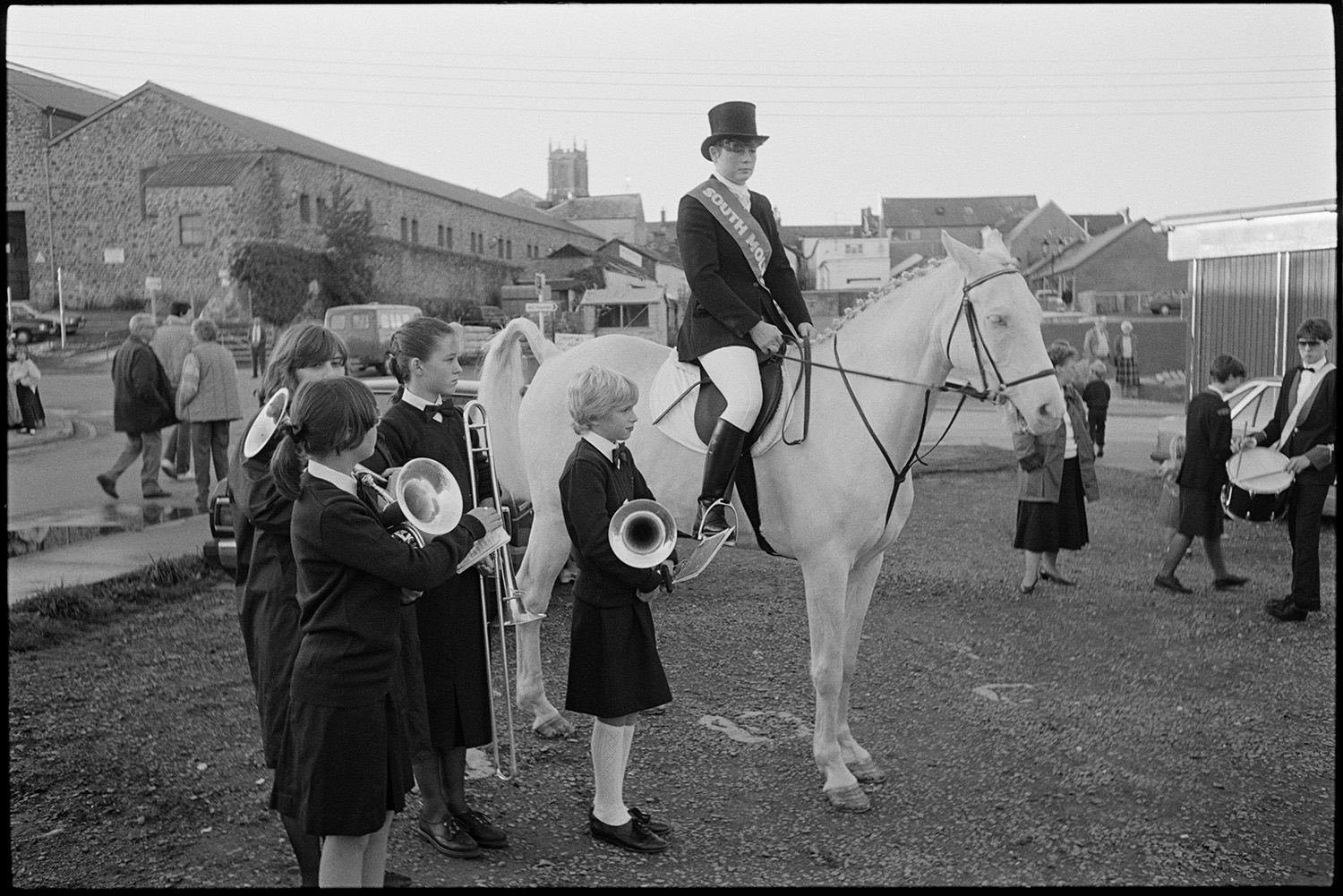 Spectators waiting for start of fair in town square. 
[Four children with instruments and a child riding a horse waiting for the start of South Molton Fair in the town square.]