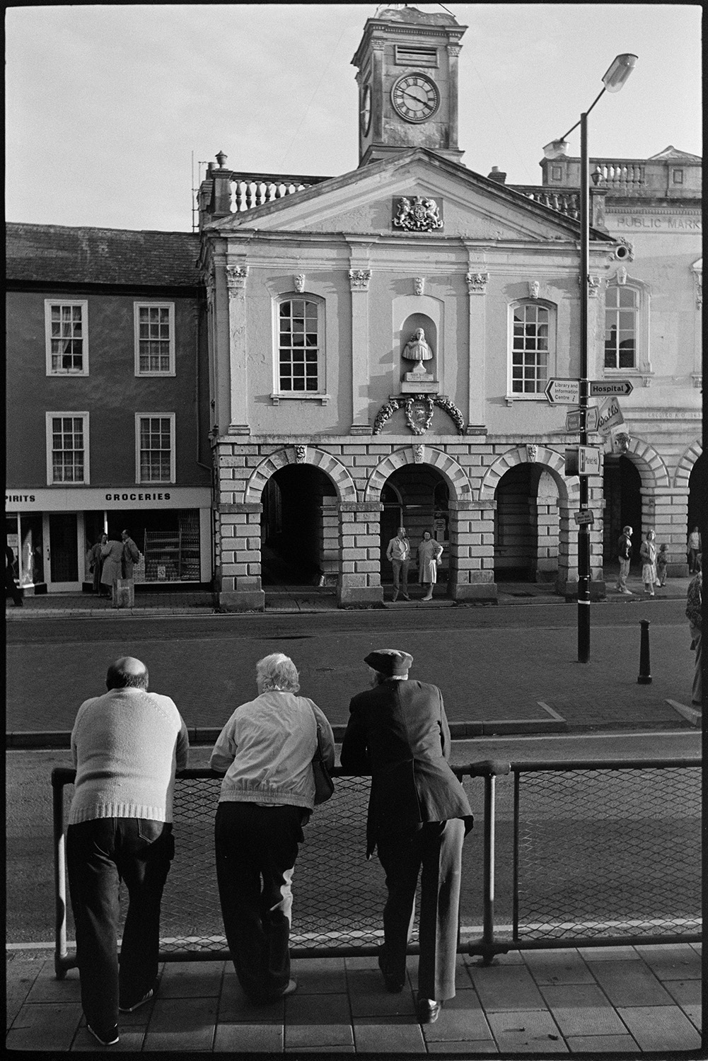 Spectators waiting for start of fair in town square. 
[Three people leaning on railings opposite South Molton Town Hall waiting for the fair to start.]