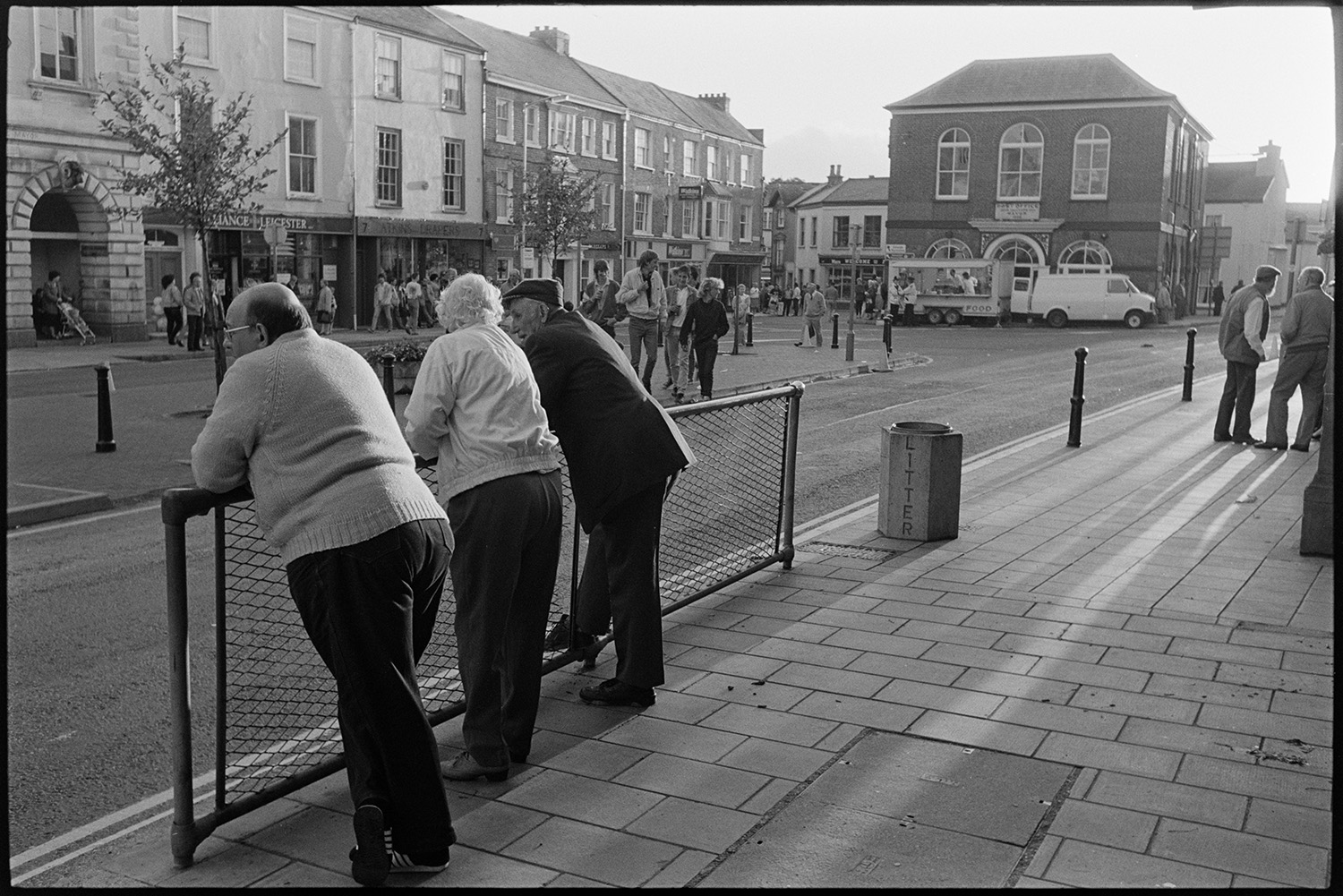 Spectators waiting for start of fair in town square. 
[Three people leaning on railings opposite South Molton Town Hall waiting for the fair to start. Other people are also gathering further along the street outside shop fronts.]