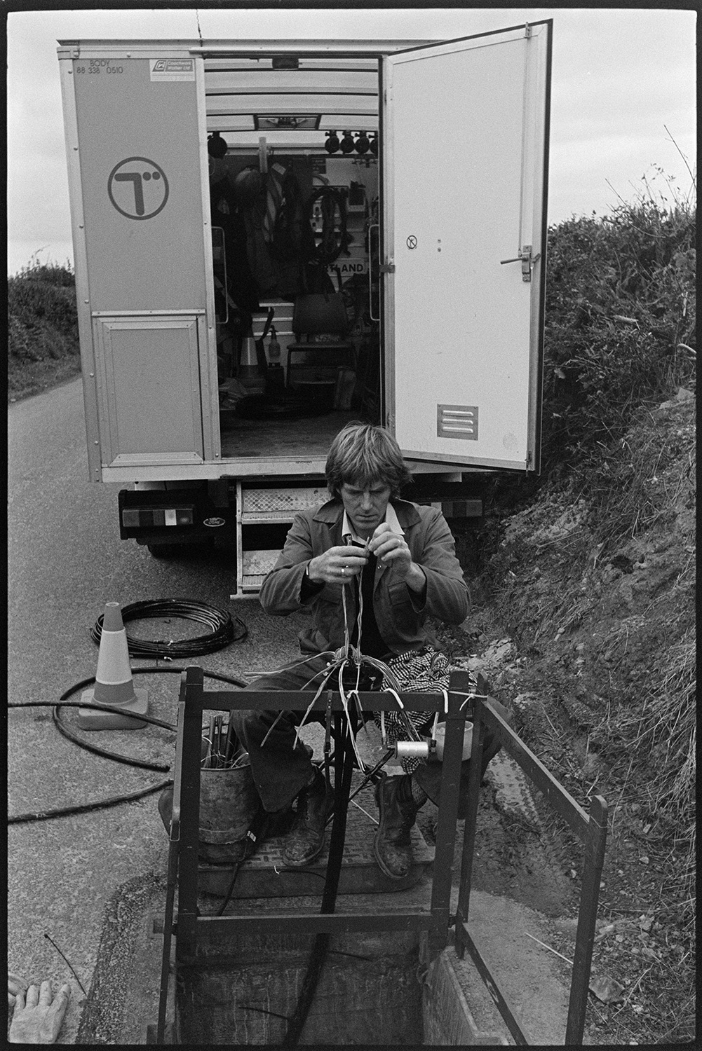 Telephone engineer working above manhole. 
[A man working on telephone cables in a manhole in a road at Ashreigney. His van is parked in the background.]
