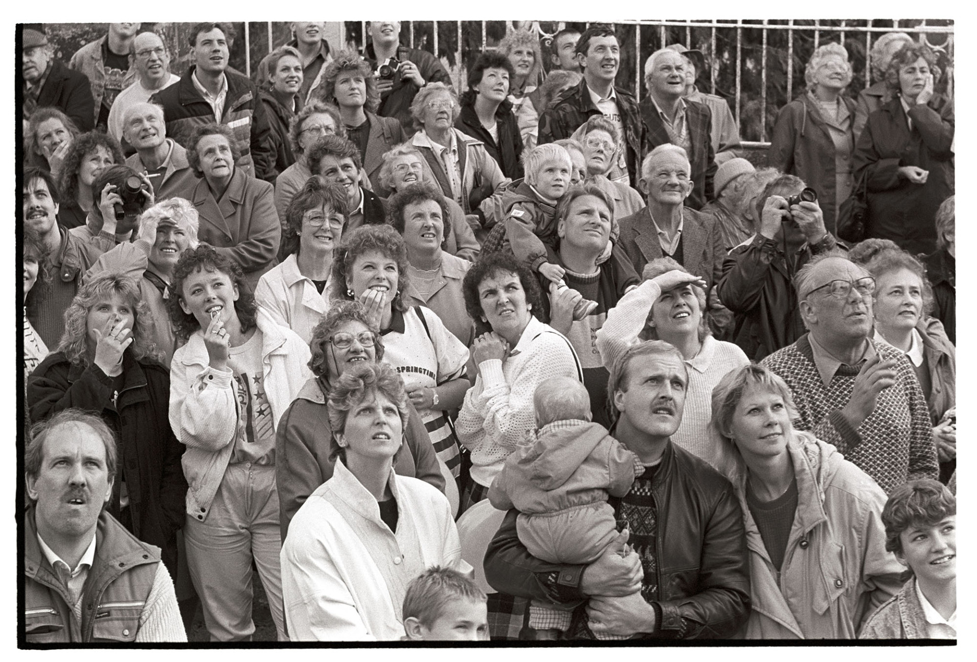 Crowd watching abseiling down church tower to raise money for restoration of the tower. 
[A crowd of men, women and children watching Barry Fewings abseil down Chulmleigh Church tower to raise money for the restoration of the tower. Two people are taking photographs. The woman wearing white in the centre is Barry Fewing's wife.]