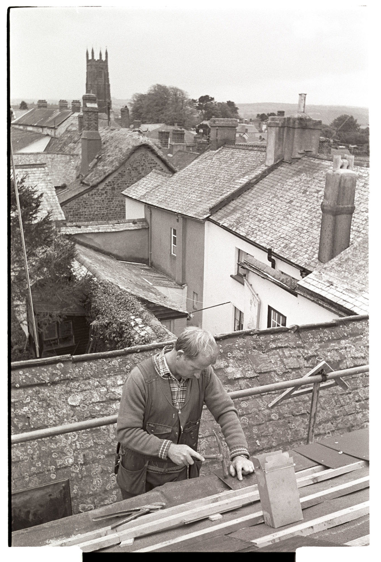 Man hanging slates with view over village with church tower. 
[Mike Hiscock hanging slates on a roof in Chulmleigh. He is stood on scaffolding and using a hammer. Roofs of other houses, both slate and thatch, and the Chulmleigh Church tower are visible in the background.]