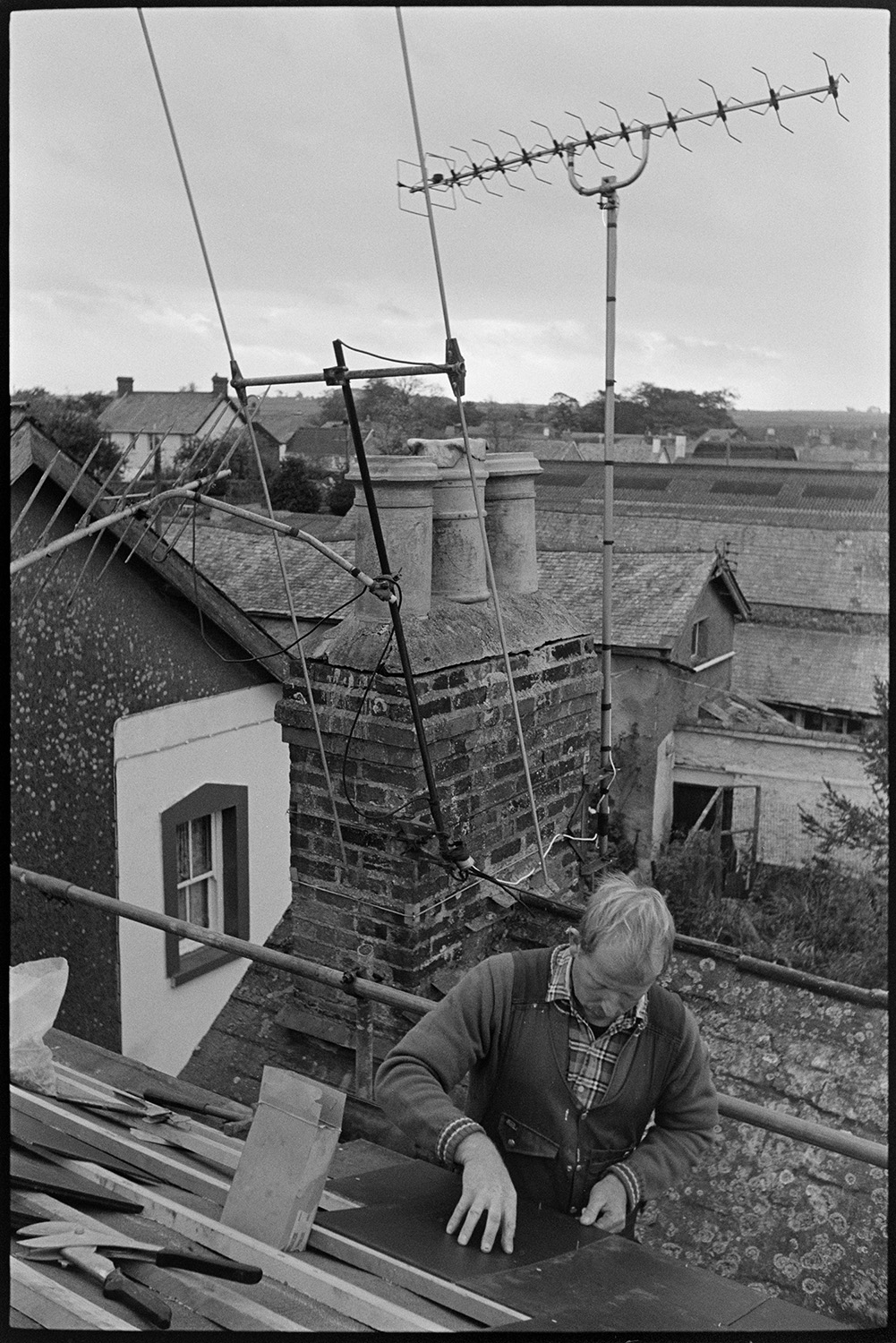 Man hanging slates with view over village with church tower. 
[Mike Hiscock hanging slates on a roof in Chulmleigh. Other rooftops and television aerials can be seen behind him.]