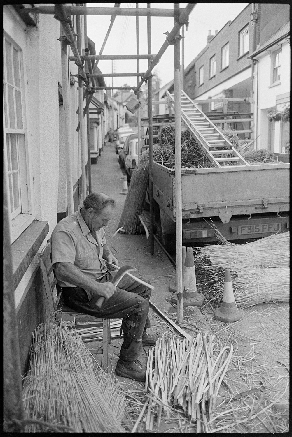 Thatcher cutting spars in village street. 
[A thatcher sat on a chair outside a house in South Molton Street, Chulmleigh, cutting spars for thatching using a billhook. Scaffolding, nitches of reed and a truck with a ladder are visible in the street.]