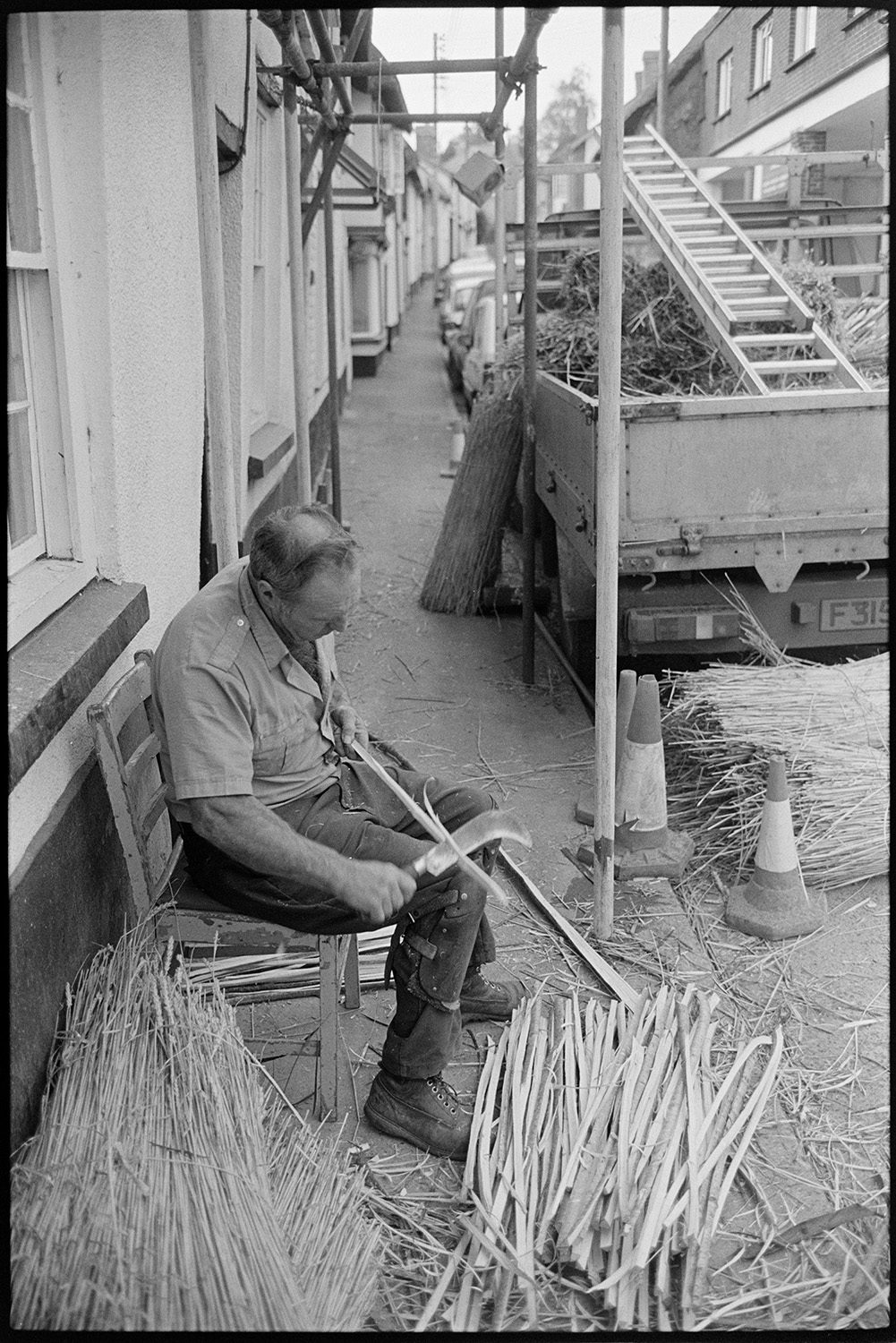 Thatcher cutting spars in village street. 
[A thatcher sat on a chair outside a house in South Molton Street, Chulmleigh, cutting spars for thatching using a billhook. Scaffolding, nitches of reed and a truck with a ladder are visible in the street.]