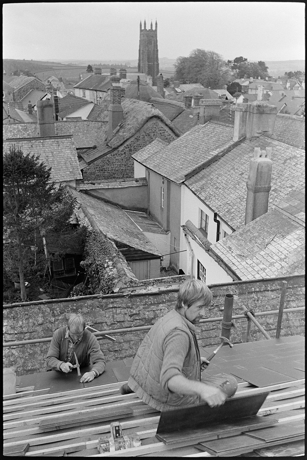 Man hanging slates with view over village with church tower. 
[Mike Hiscock, in the background, and another man hanging slates on a roof in Chulmleigh. Other rooftops and the church tower can be seen in the background.]