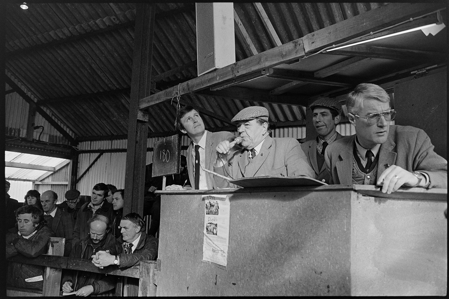 Ring at cattle market, farmers bidding, auctioneers in box. 
[Auctioneers in a box by the ring at a cattle auction near Wheddon Cross. One of them is speaking into a microphone.]