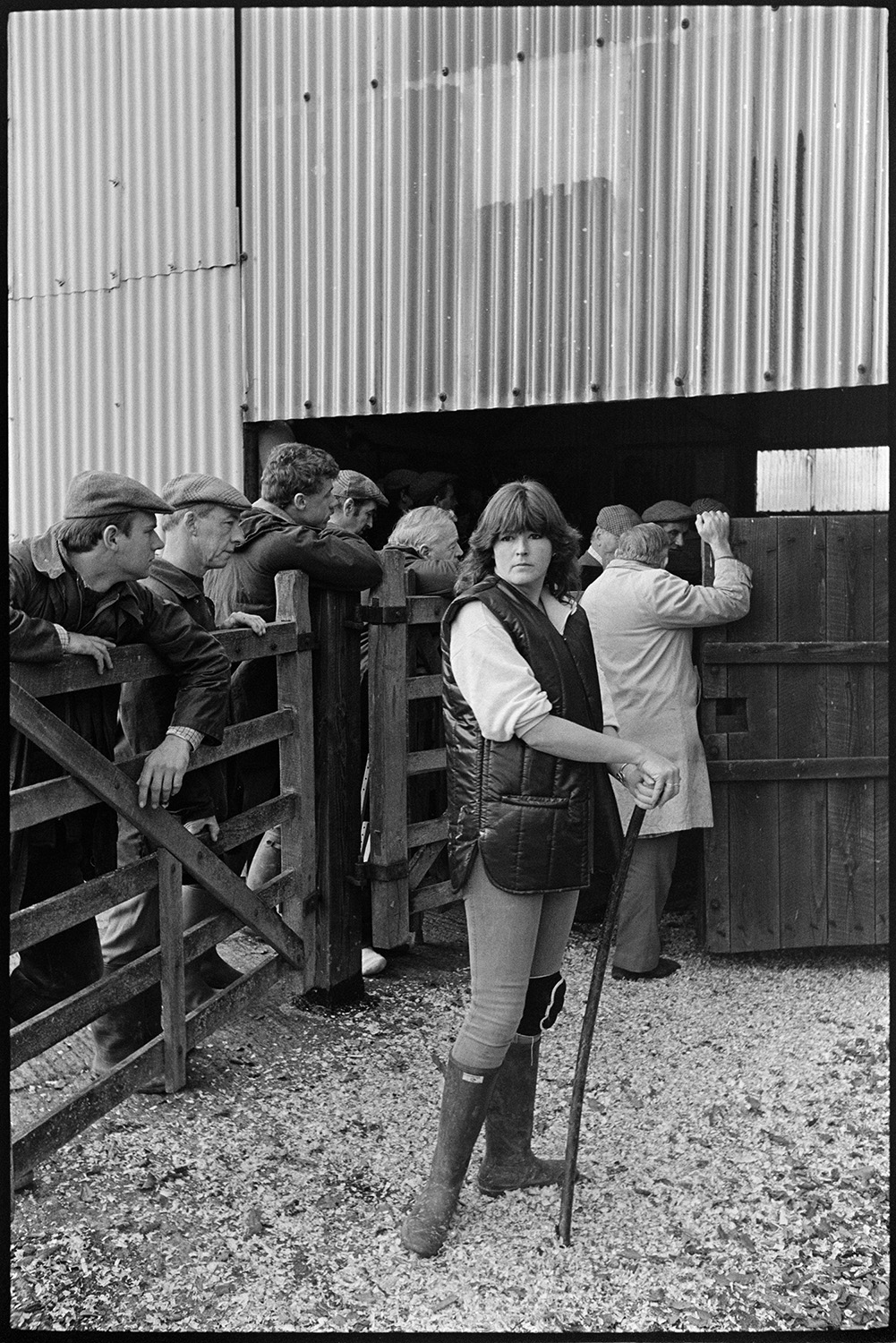 Ring at cattle market, farmers bidding, auctioneers in box. 
[A woman working at a cattle market near Wheddon Cross. Men are looking over a wooden fence in the background to the cattle ring.]