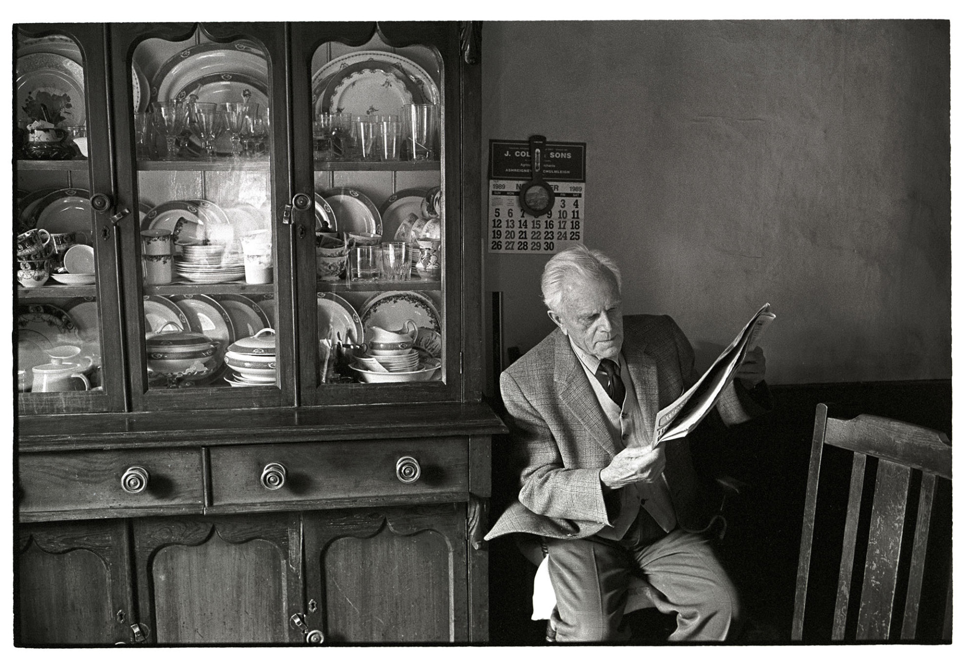 Man reading seated next to dresser with china, Dresser made in Lapford. 
[Ron Jury sat reading a newspaper next to a wooden dresser filled with china, in a house at Leigh Road, Chulmleigh. A calendar is hung on the wall behind him. The dresser was made in Lapford.]