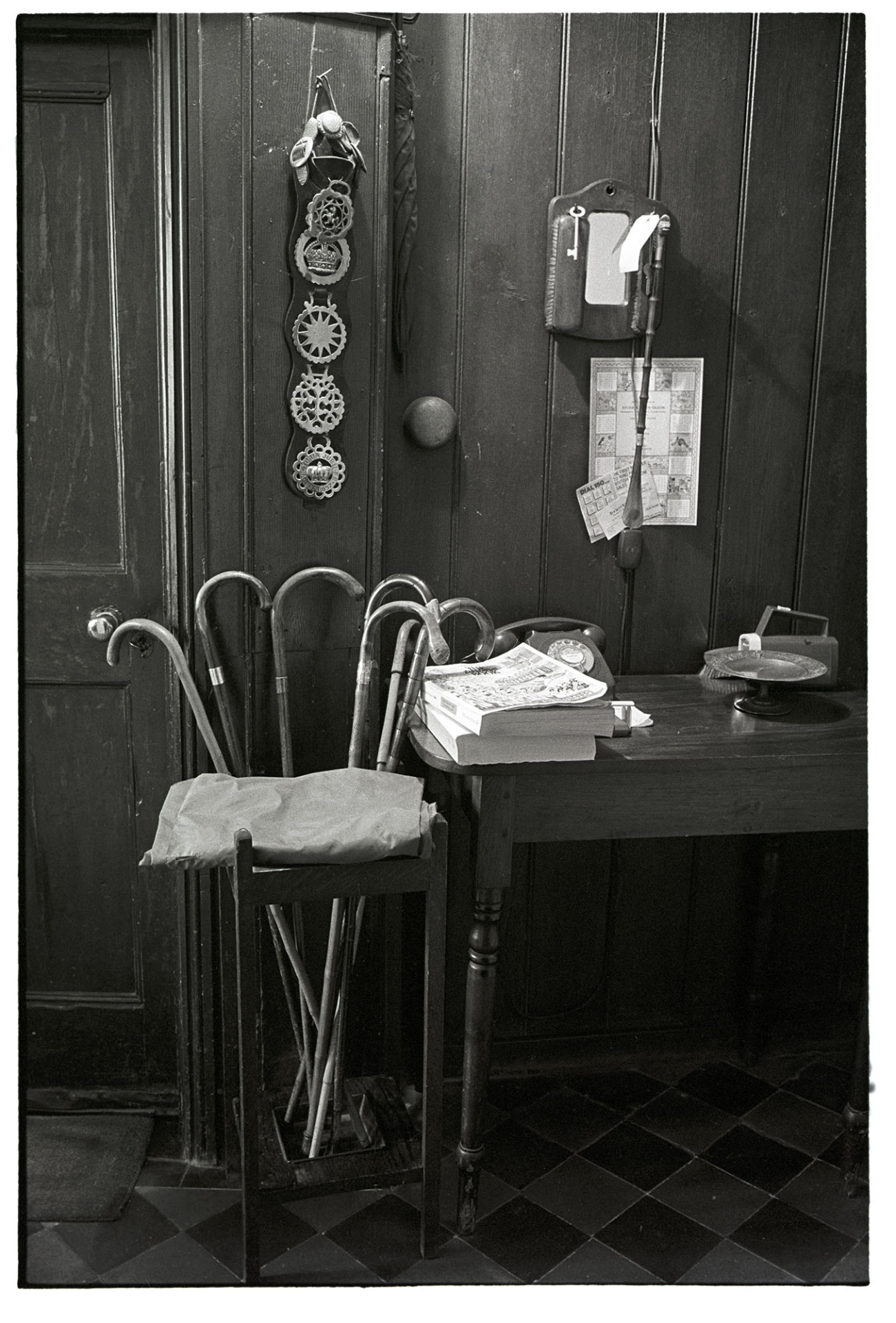 Panelled hall with telephone table and walking sticks, horse brasses on wall. 
[A wooden panelled hallway at a house in Leigh Road Chulmleigh. A desk with a telephone and telephone directory, walking stick stand with walking sticks and horse brasses hung on the wall are all in the hallway.]