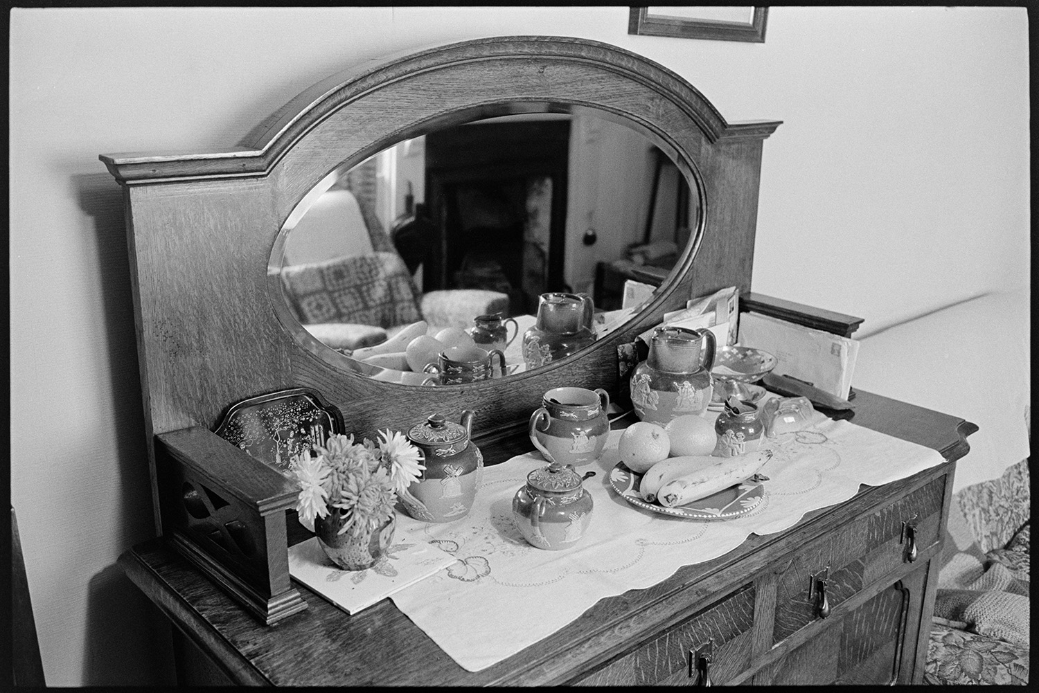 Sideboard with mirror and china in front room. 
[A sideboard with a mirror, china teapot and jug, vase of flowers, tray of fruit and post, all laid on a cloth in the front room of a house in Leigh Road, Chulmleigh.]