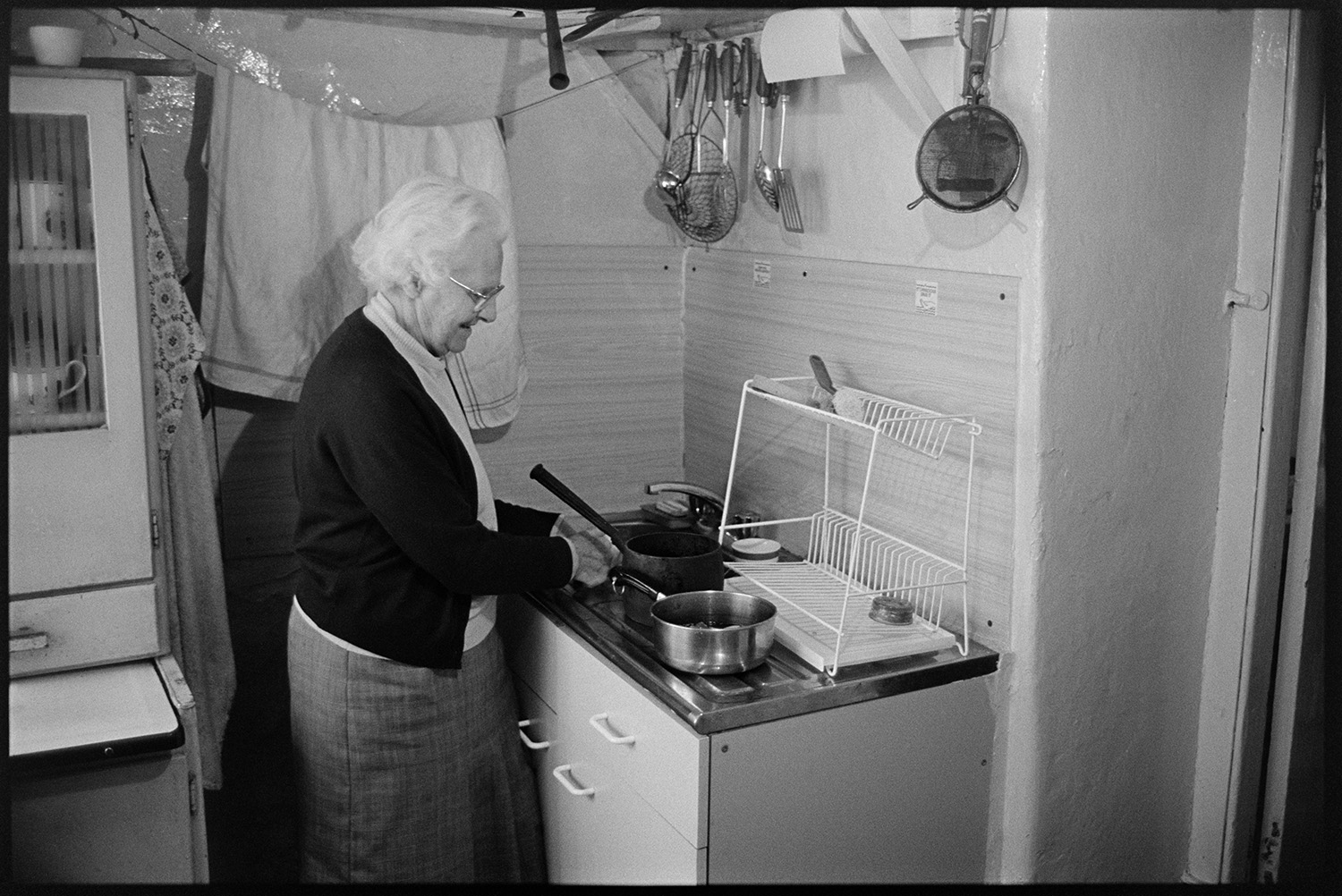 Woman working at kitchen sink. 
[Winnie Jury washing saucepans in her kitchen sink in her house at Leigh Road, Chulmleigh. A rack is visible on the draining board and cooking utensils are hung above the sink.]