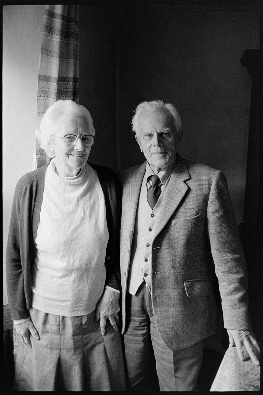 Man and woman standing at window. 
[Ron Jury and Winnie Jury standing by a window in their house at leigh Road, Chulmleigh.]