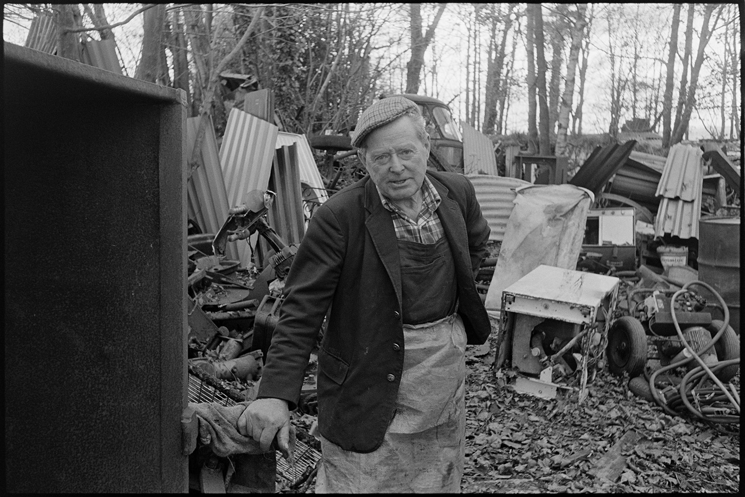 Scrap metal merchant working in his yard, chatting to photographer. 
[Jimmy Hughes in his scrap metal yard at Leigh Road, Chulmleigh, facing the camera. Various items can be seen I the yard, including sheets of corrugated iron, old engine parts and an old washing machine.]
