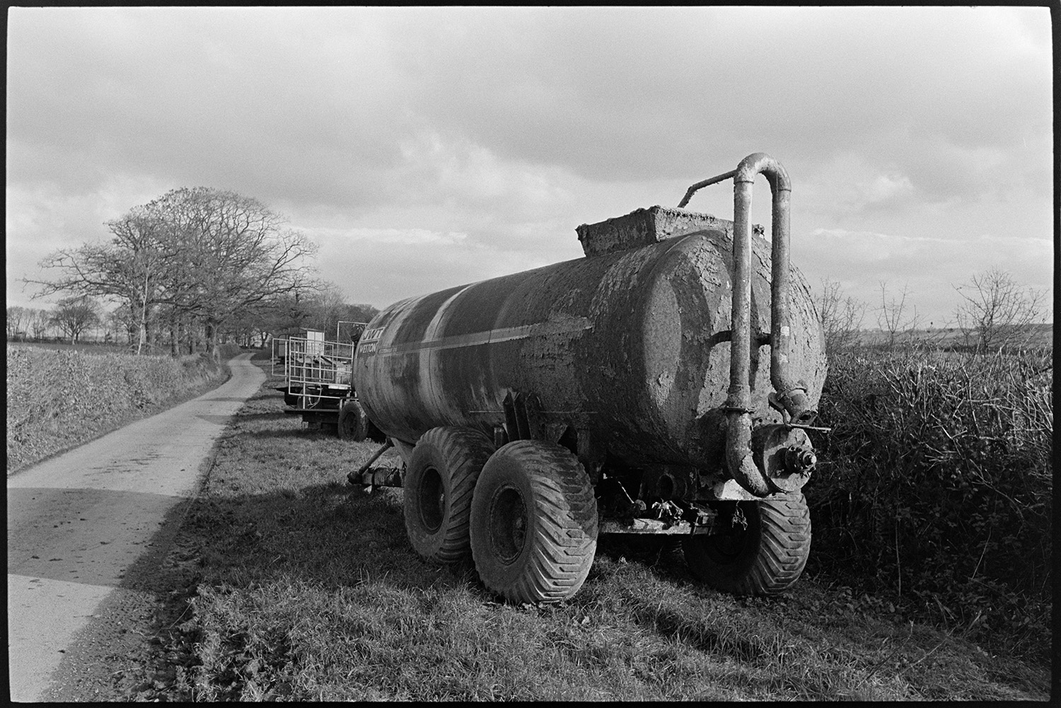 Large silage or muck tanker parked beside road. 
[A muck spreader or silage tanker parked on the verge between a hedge and road at Parsonage Farm, Chulmleigh. Other trailers are parked further along the verge.]