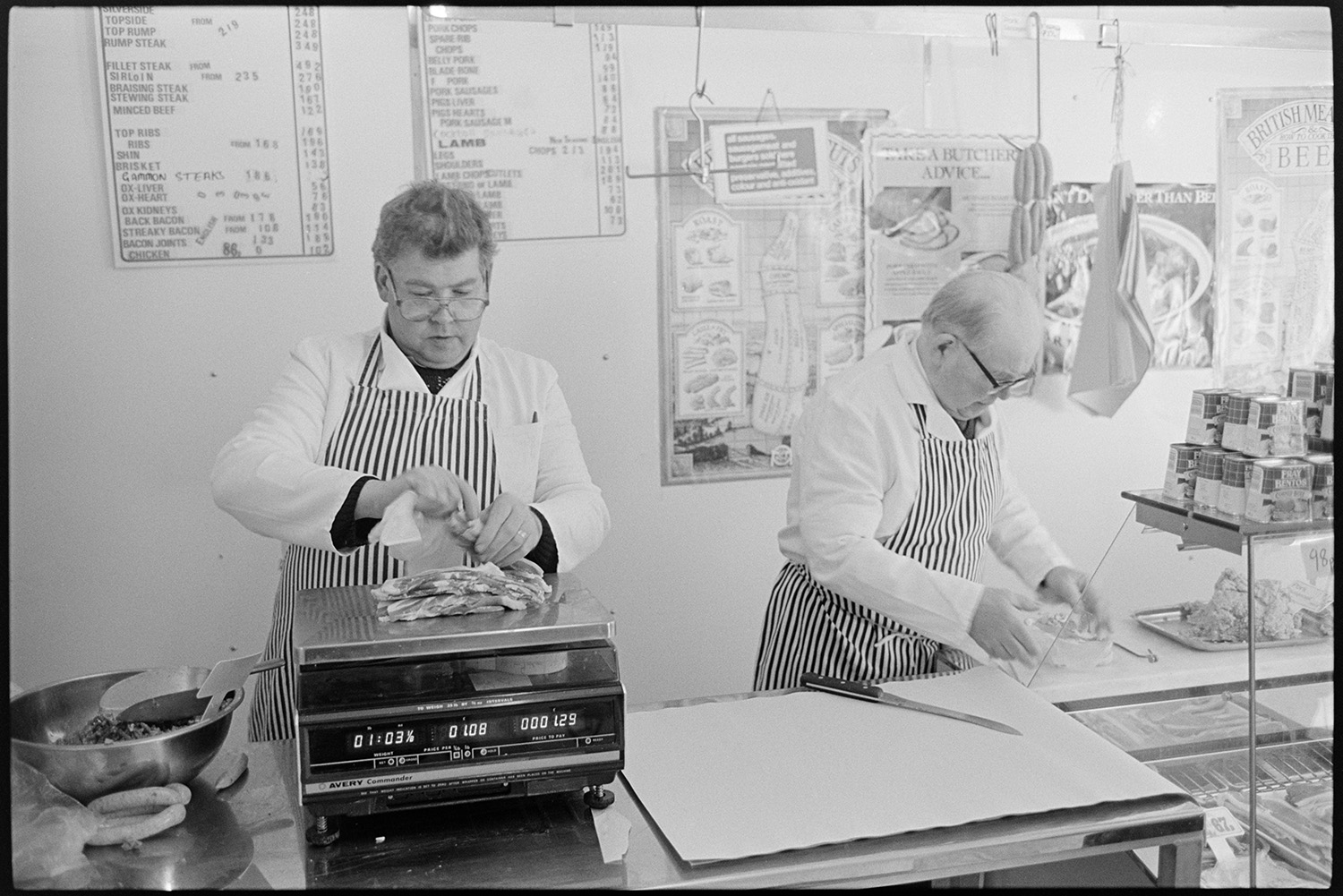Butchers shop, proprietor and customers, cash desk, butcher weighing meat. 
[Two butchers preparing meat on the counter of W Cann butcher's shop in South Molton Street, Chulmleigh. One of them is weighing meat. Posters and a price list are displayed behind the counter.]