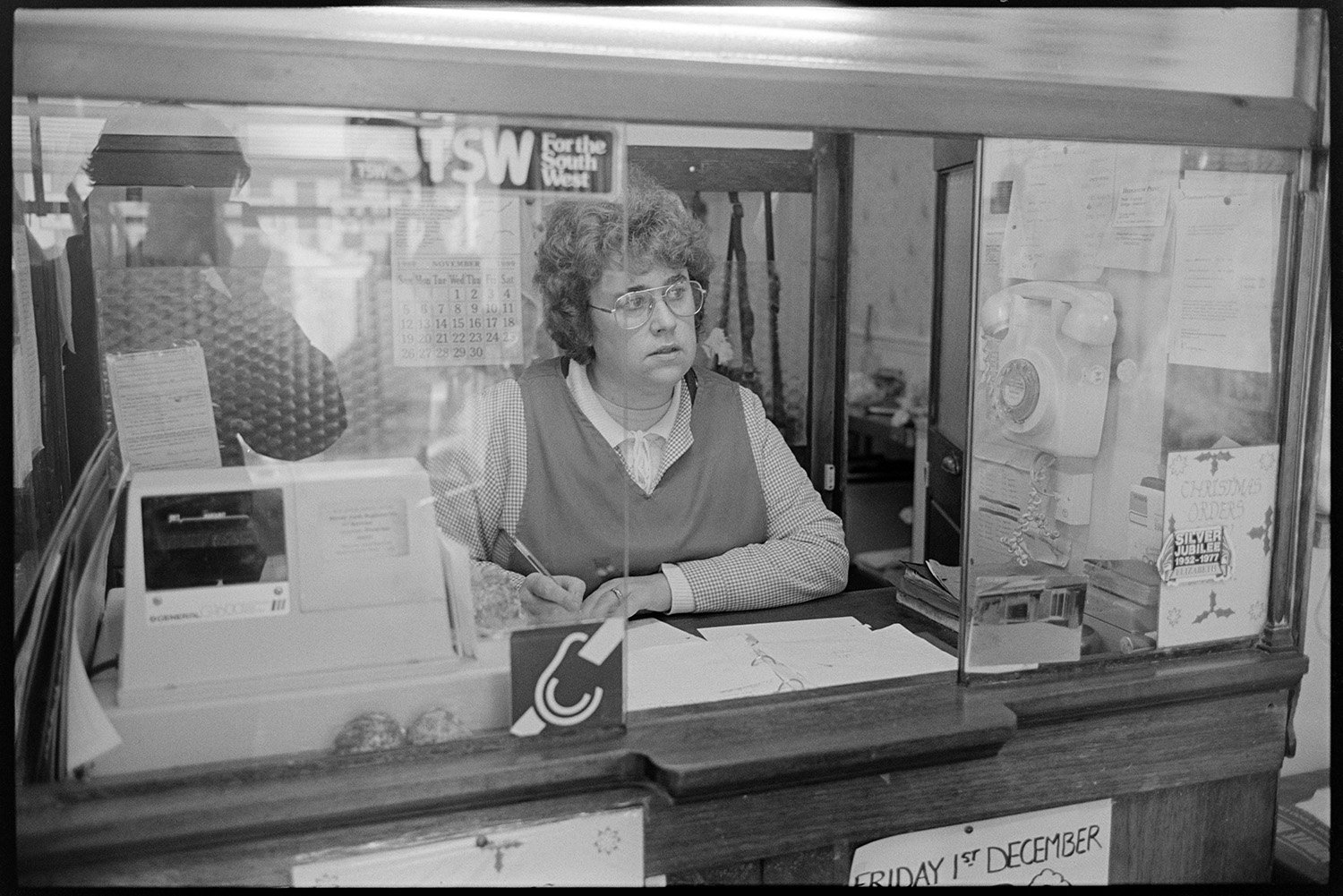 Butchers shop, proprietor and customers, cash desk, butcher weighing meat. 
[A woman at the till kiosk in W Cann butcher's shop in South Molton Street, Chulmleigh. She is writing in a notebook by the cash register. A telephone is fixed to the wall.]