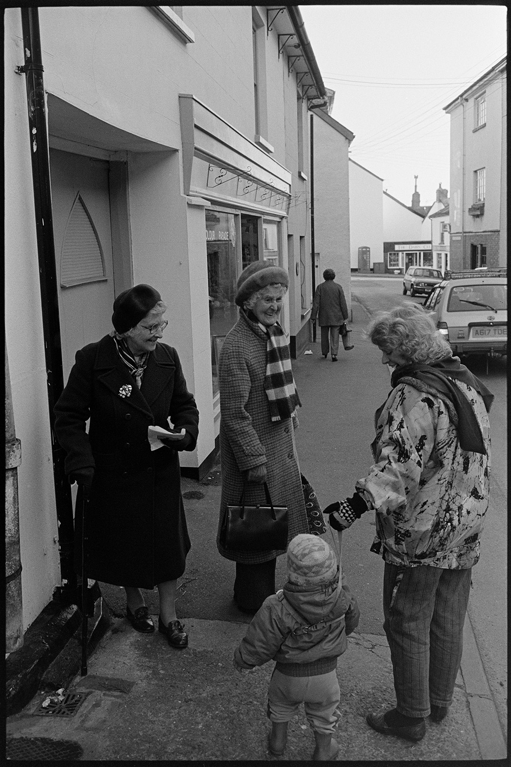 Street scenes, women and children. Clothes, coats, woolly hats. 
[A woman and child talking to two older women near a shop front window in South Molton Street, Chulmleigh. They are wearing coats, hats, scarves and gloves.]