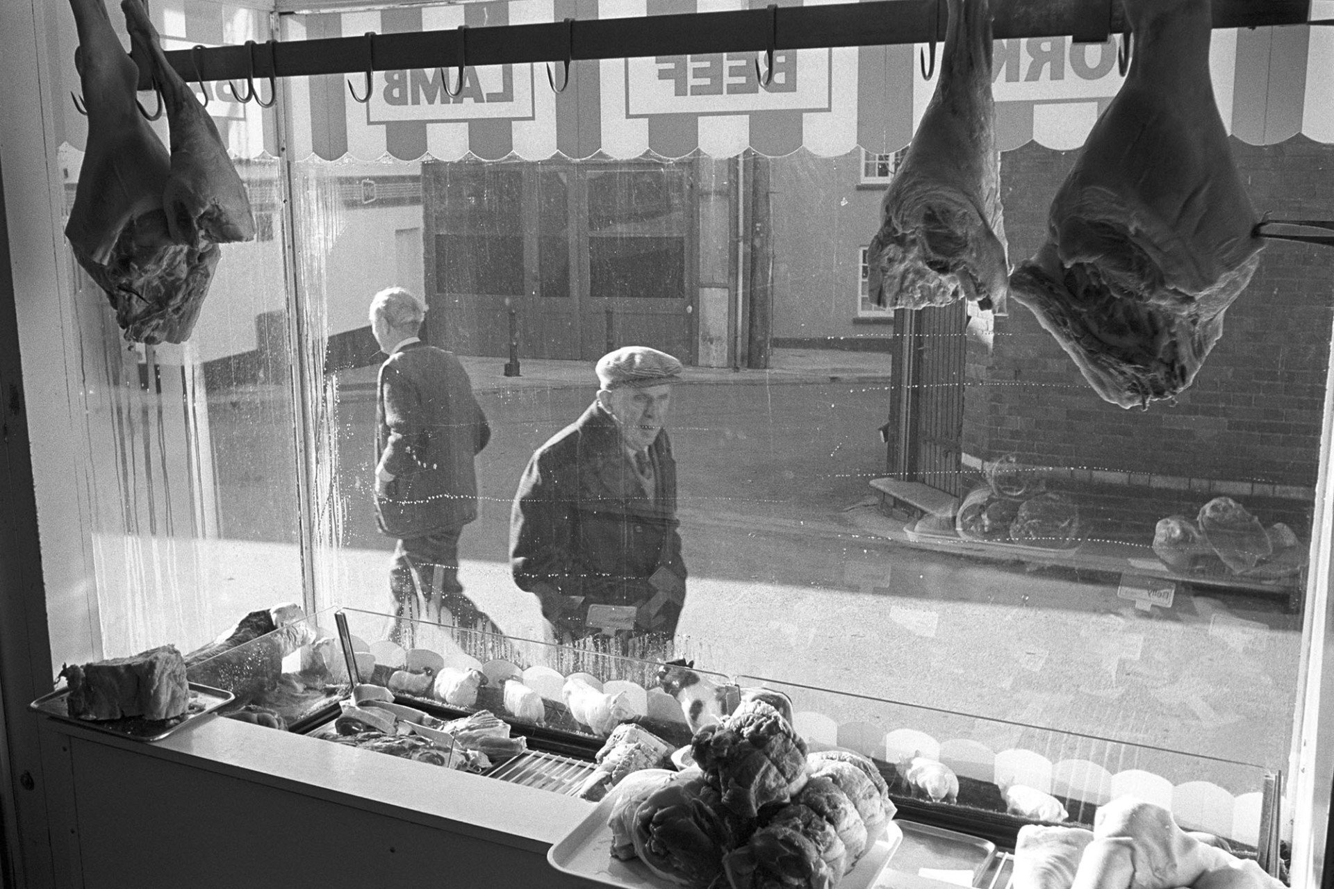 Window of butchers shop with passers by, seen from inside. 
[Cuts of meat hanging up in Cann's Butcher's shop window on South Molton Street, Chulmleigh. The image is taken from inside the butcher's shop and two men can be seen walking past outside.]