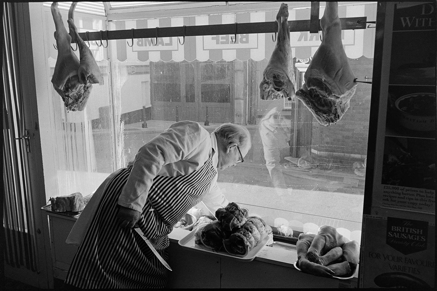 Window of butchers shop with passers by, seen from inside, butcher selecting meat. 
[A butcher selecting cuts of meat from the window display in W Cann butchers shop in South Molton Street, Chulmleigh.]
