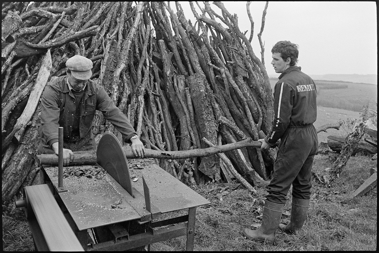 Farmers cutting logs, circular saw, woodpile. 
[Two men cutting up logs using a circular saw in a field at Parsonage Farm, Chulmleigh. A woodpile is stacked up behind them.]