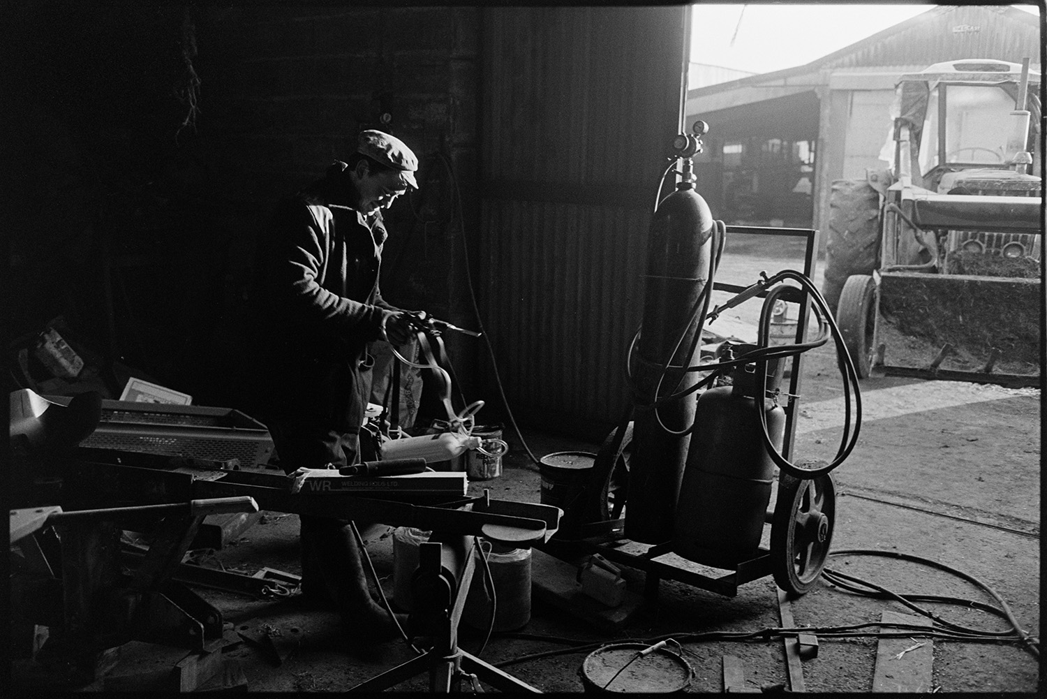 Farmer preparing welding torch in shed. 
[Bill Crocombe preparing a welding torch in a barn at Parsonage Farm, Chulmleigh. Two gas canisters are on a sack truck next to him and a tractor is parked in front of another barn in the farmyard.]