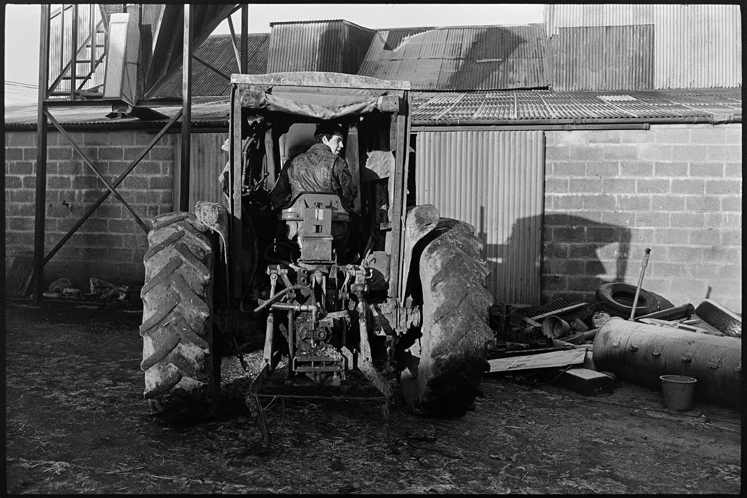 Farmyard scenes, bullocks, sheep, silage under polythene. 
[A man driving a tractor in the farmyard at Parsonage Farm, Chulmleigh. He is parked in front of a brick and corrugated iron barn with various items piled outside.]