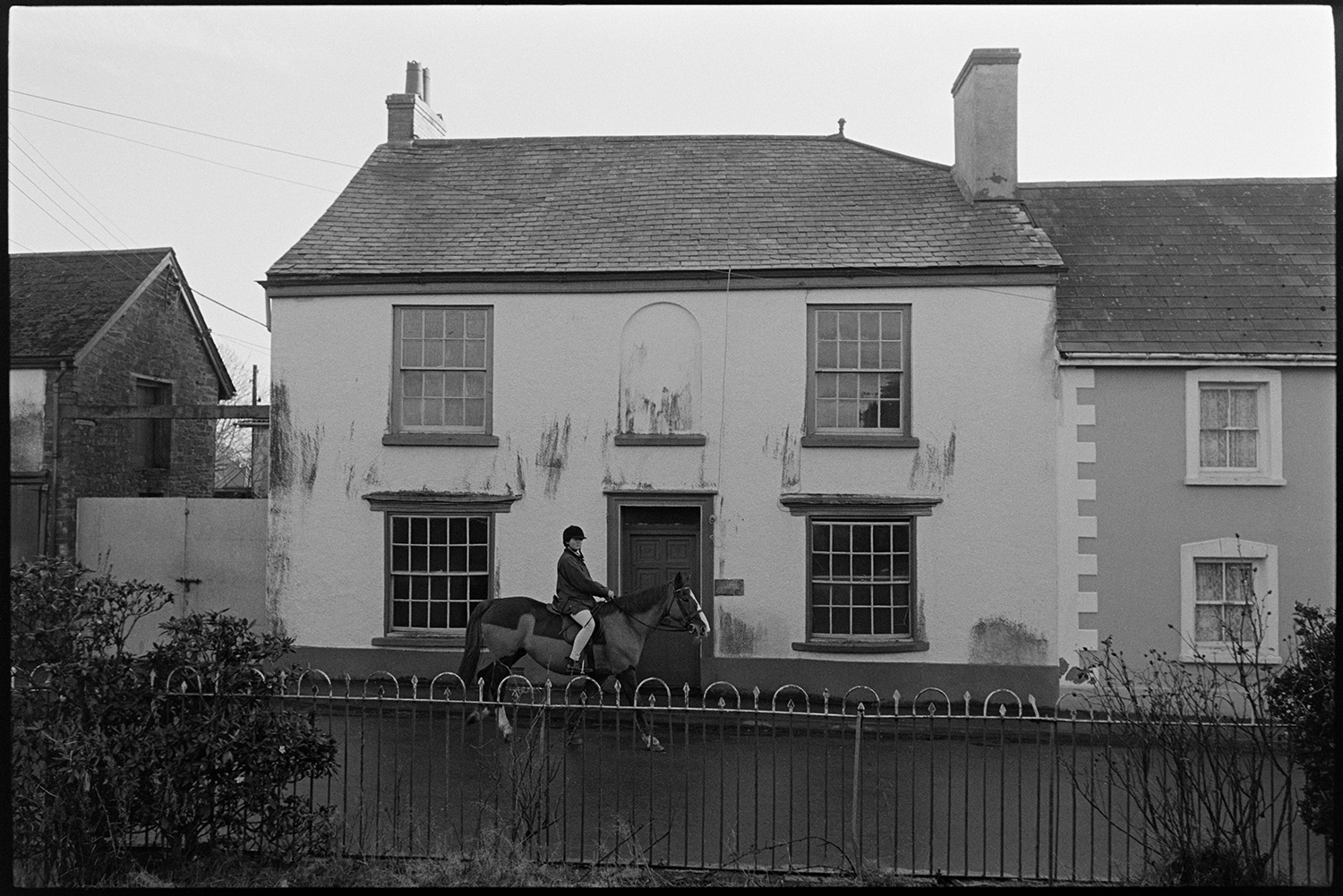 Street scenes. 
[A mounted horse rider, riding along a street in Chulmleigh past a house. Metal railings are visible in the foreground.]