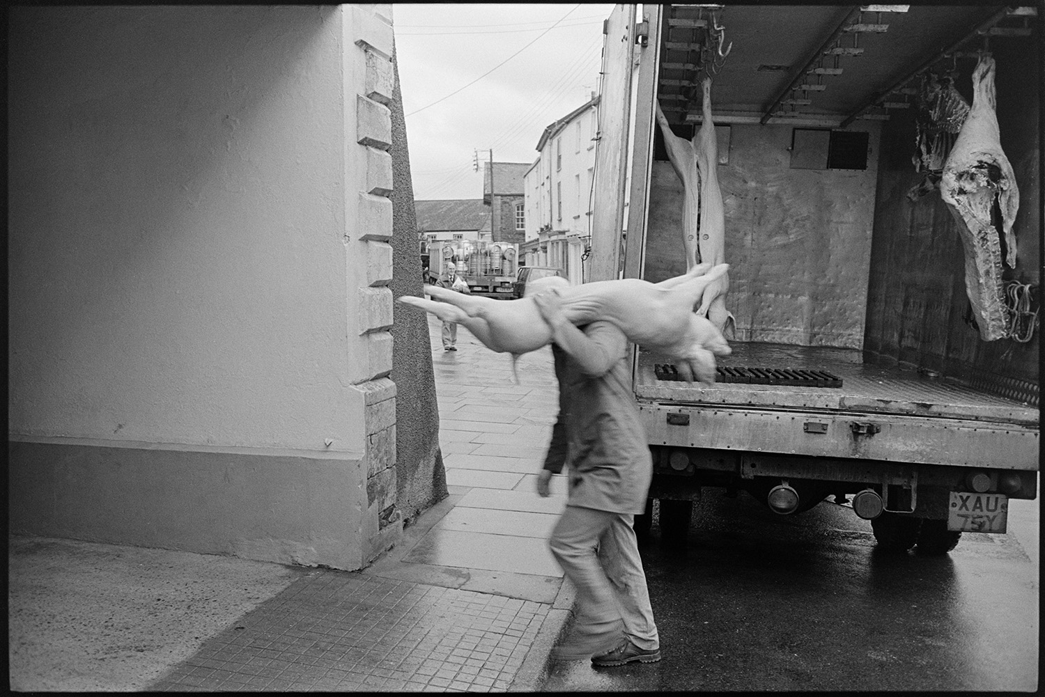 Butcher unloading carcasses of meat from lorry. 
[A butcher carrying a pig carcase from a meat lorry into his shop in South Molton Street, Chulmleigh. Other carcasses and cuts of meat are hung up in the lorry.]