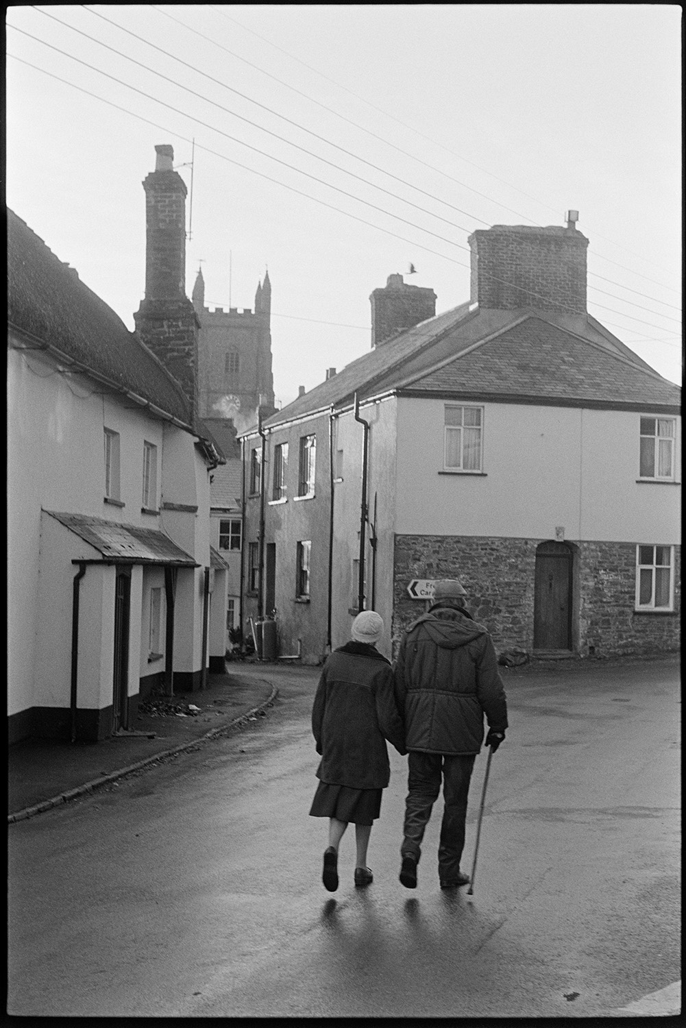 Street scenes. 
[A man and woman walking along a street in Chulmleigh holding hands. The man is using a walking stick. They are walking past houses. The church tower can be seen in the background.]