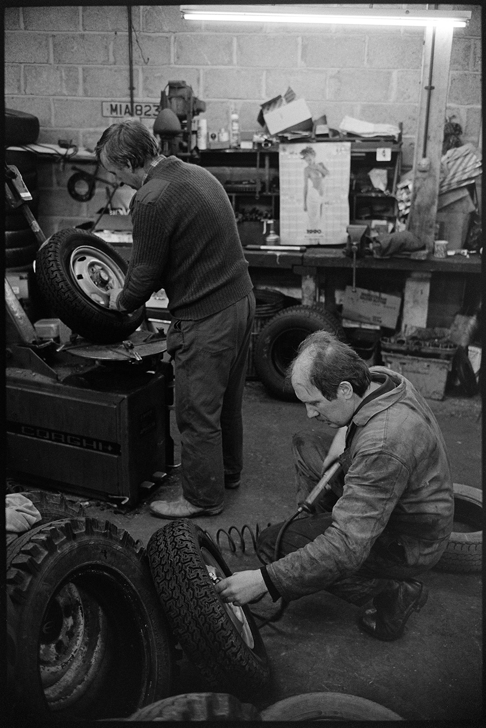 Garage workshop, man welding, chatting in office, general view of premises. 
[Winston Pincombe, standing, and another man checking tyres in the workshop at the Winston Pincombe Garage in South Molton Street, Chulmleigh.]
