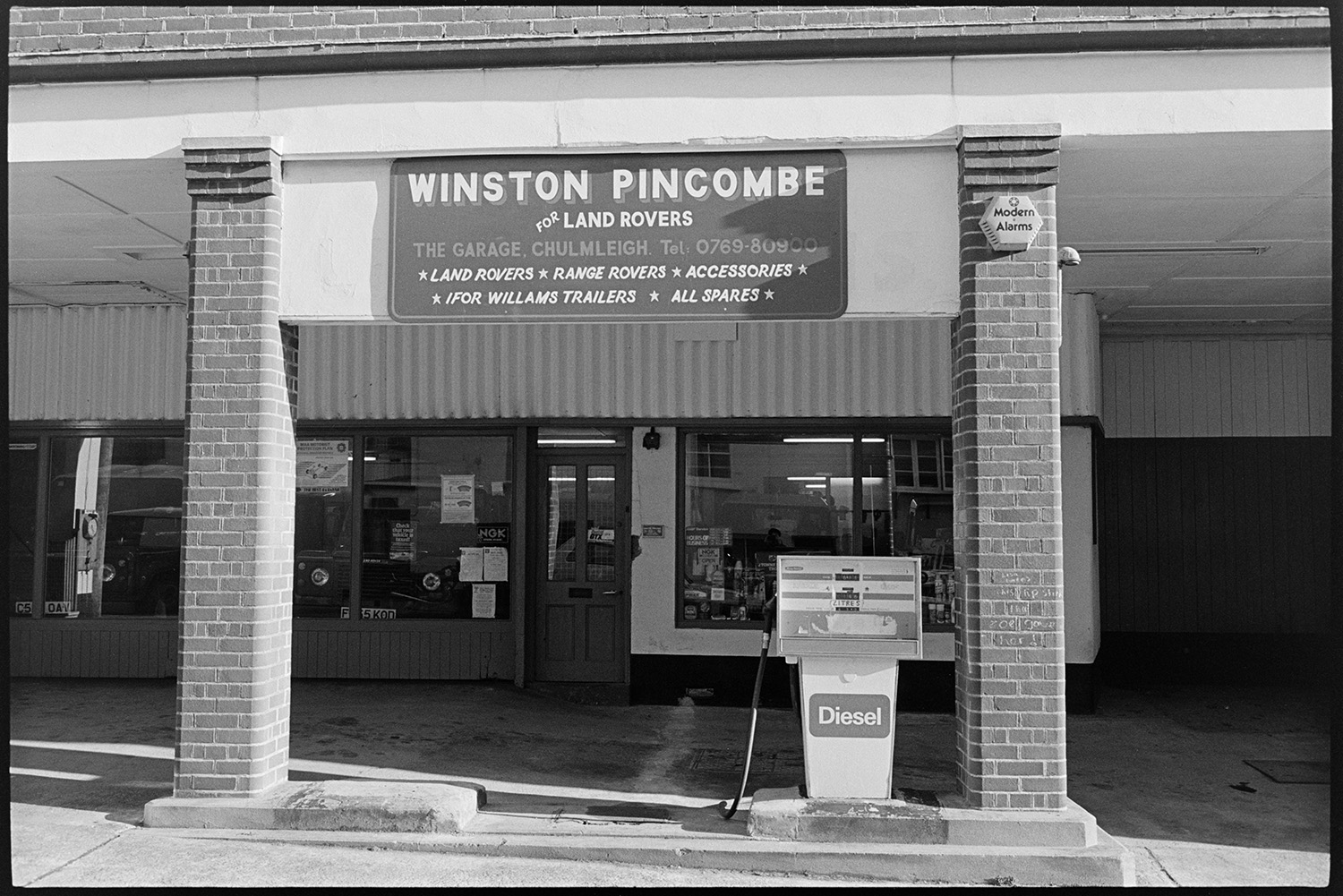 Garage workshop, mechanics at work, proprietor in office, front of premises, store room. 
[The front of the Winston Pincombe Garage in South Molton Street, Chulmleigh. The garage sign and diesel pump are visible outside.]