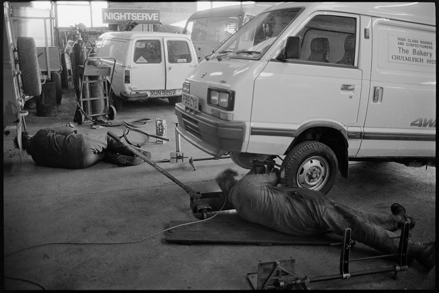 Garage workshop, mechanics at work, proprietor chatting man in store room, Land Rover. 
[Two mechanics working on a Land Rover and a van at the Winston Pincombe Garage in South Molton Street, Chulmleigh. More vans can be seen in the background.]
