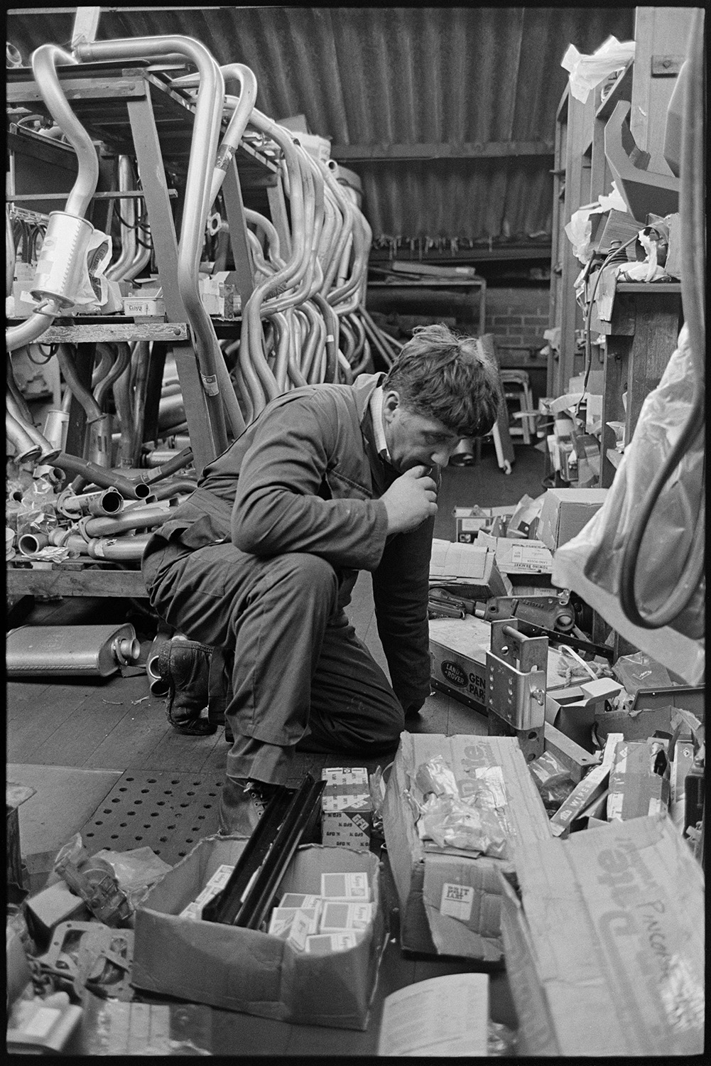 Garage workshop, mechanics at work, proprietor chatting man in store room, Land Rover. 
[A man looking at items in boxes in the store room at the Winston Pincombe Garage in South Molton Street, Chulmleigh. Various pipes can be seen in the background.]