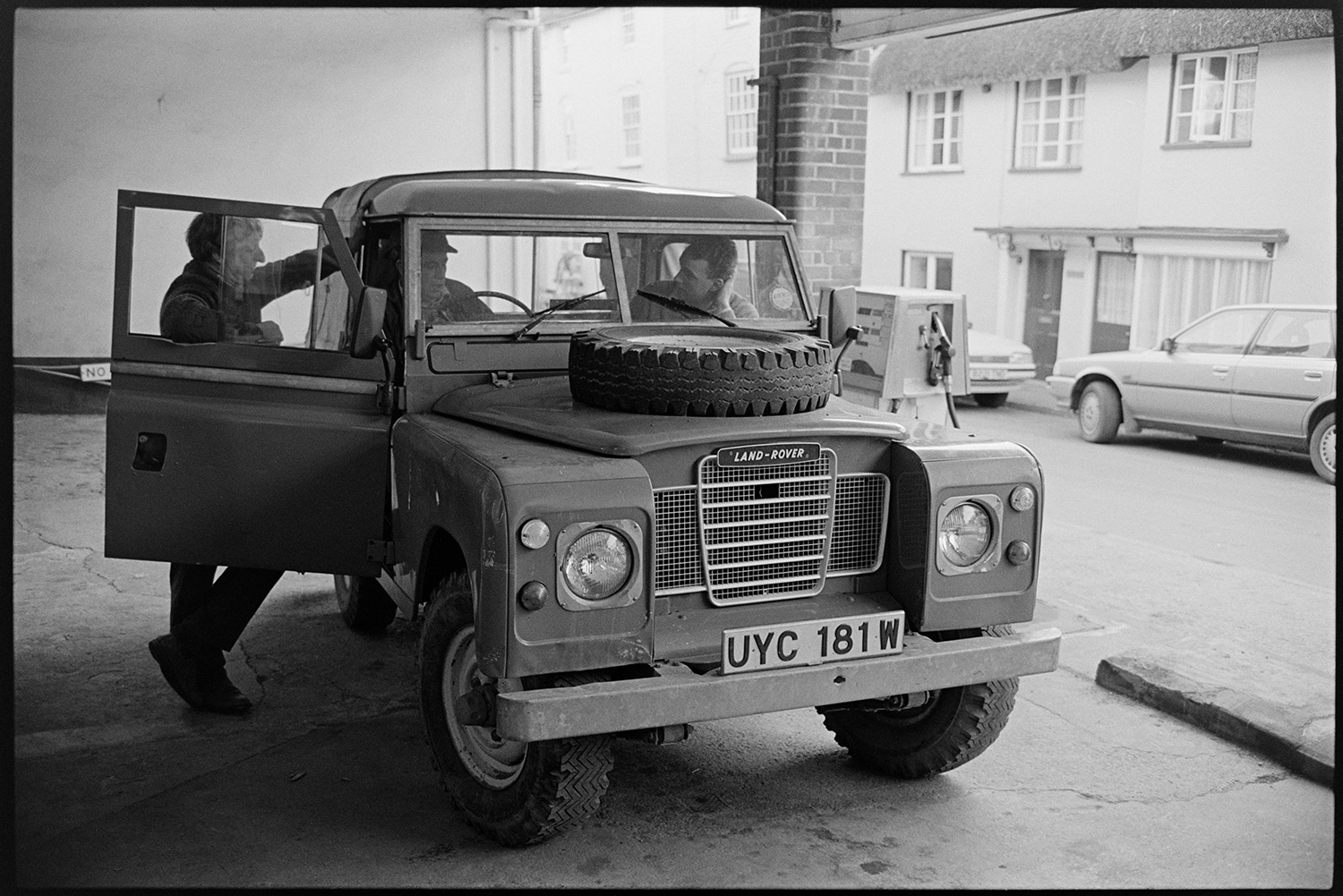 Garage workshop, mechanics at work, proprietor chatting man in store room, Land Rover. 
[Three men talking by a Land Rover at the Winston Pincombe garage in South Molton Street in Chulmleigh. Two of the men are sat inside the Land Rover.]