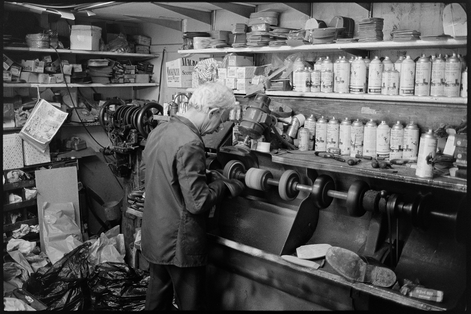 Man and woman shoemakers working in shoe repair shop. 
[Ray Gale working on the sole of a shoe in his shoe repair shop in Boutport Street, Barnstaple. A spindle with various wheels is turning in front of him and aerosol cans are on the shelves in the background.]