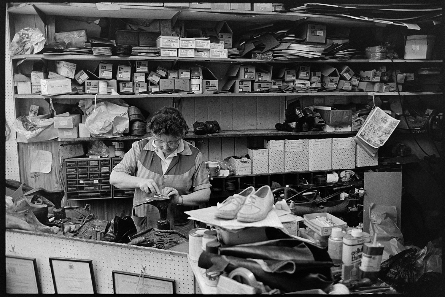 Man and woman shoemakers working in shoe repair shop. 
[Mrs Gale working on a shoe repair on top of a shoe mould in her shoe repair shop in Boutport Street, Barnstaple. Shoe boxes can be seen stacked on shelves in the background.]